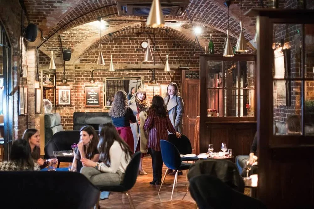 Women engaging in conversation inside a cozy brick-walled restaurant or cafe, with tables set for dining and warm lighting.
