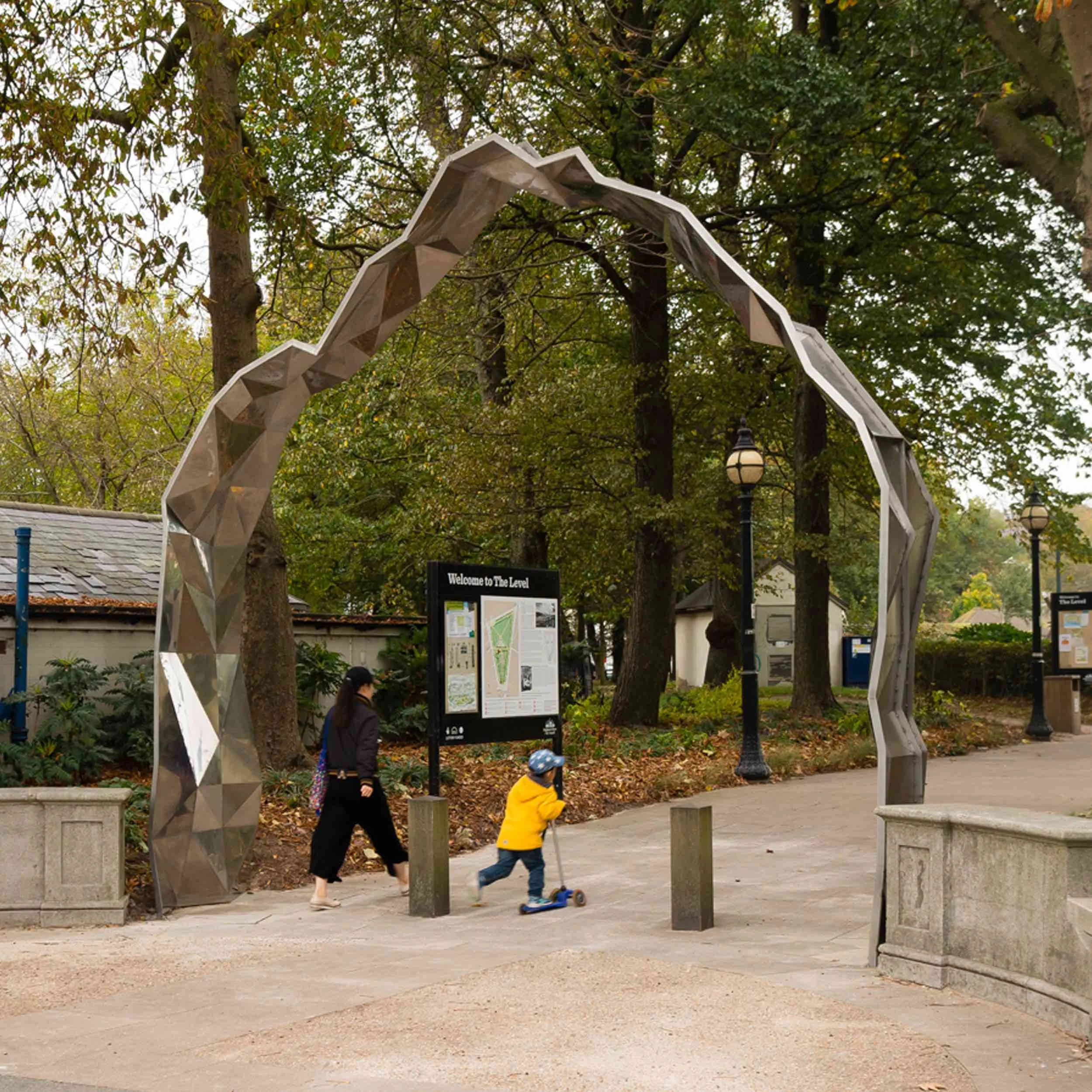 A park entrance archway made of reflective metal with geometric faceted design. Two people, a woman and a young child, walk and ride a scooter near the entrance, surrounded by trees and lampposts. An informational sign is visible behind them.
