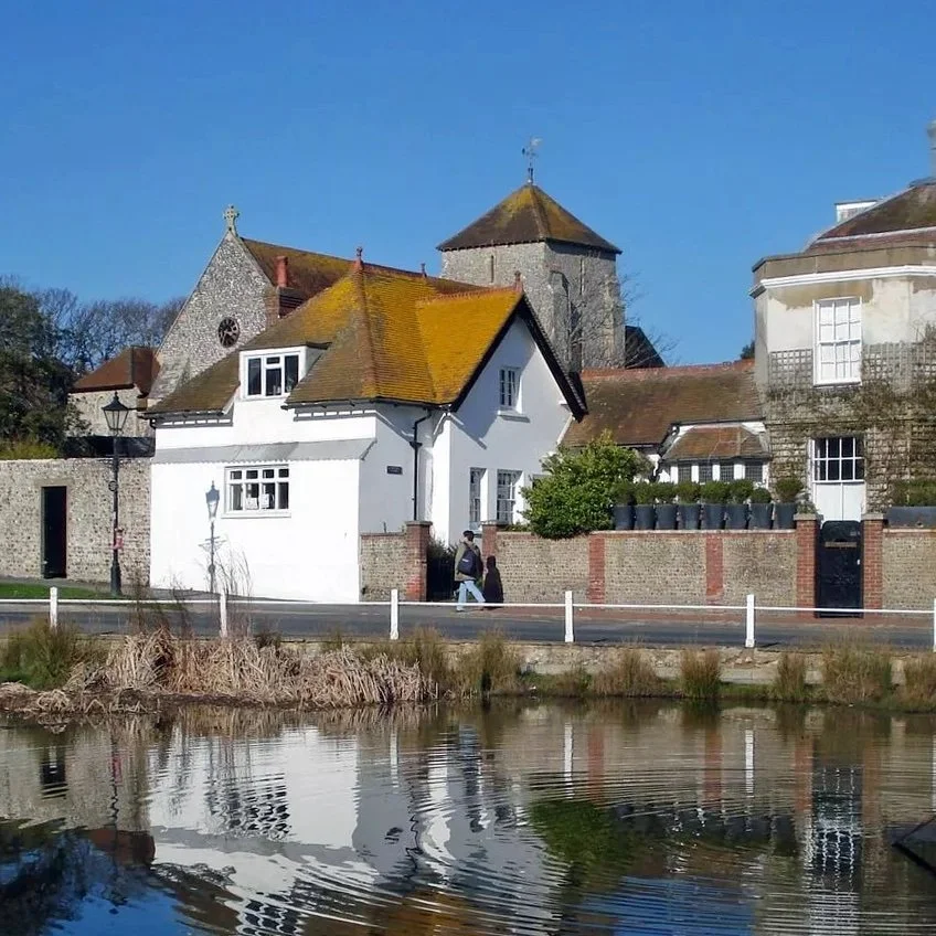 Historic European-style buildings along a canal with reflections in the water, clear blue sky, and some pedestrians walking.