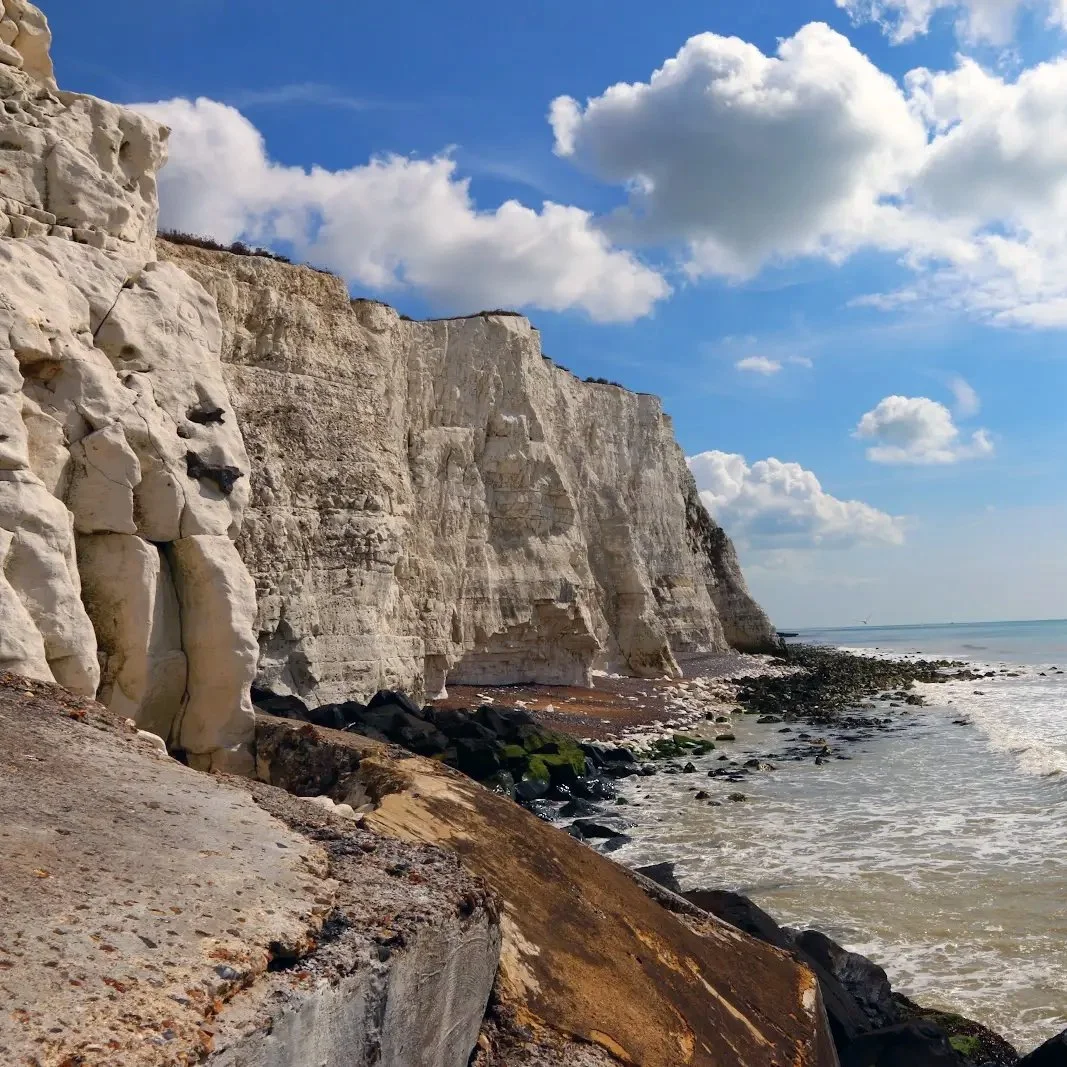 White chalk cliffs alongside a rocky beach on a sunny day with blue sky and scattered clouds.