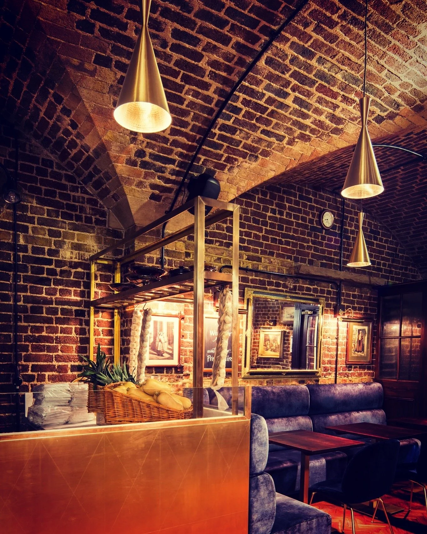Interior of a cozy restaurant with a brick vaulted ceiling, pendant lights, framed art on brick walls, a blue velvet banquette, and a metal and wood partition with a basket of bread and garlic.