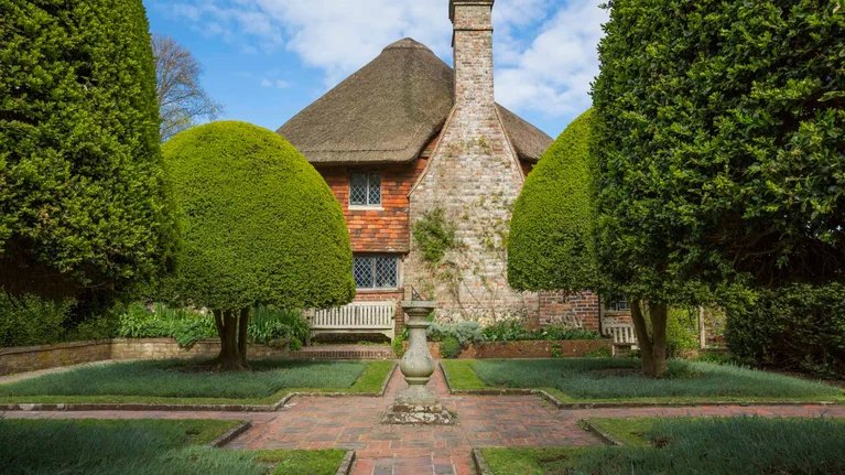 A brick house with a steep thatched roof and a stone chimney, surrounded by a well-manicured garden with trimmed bushes and a fountain in the center.