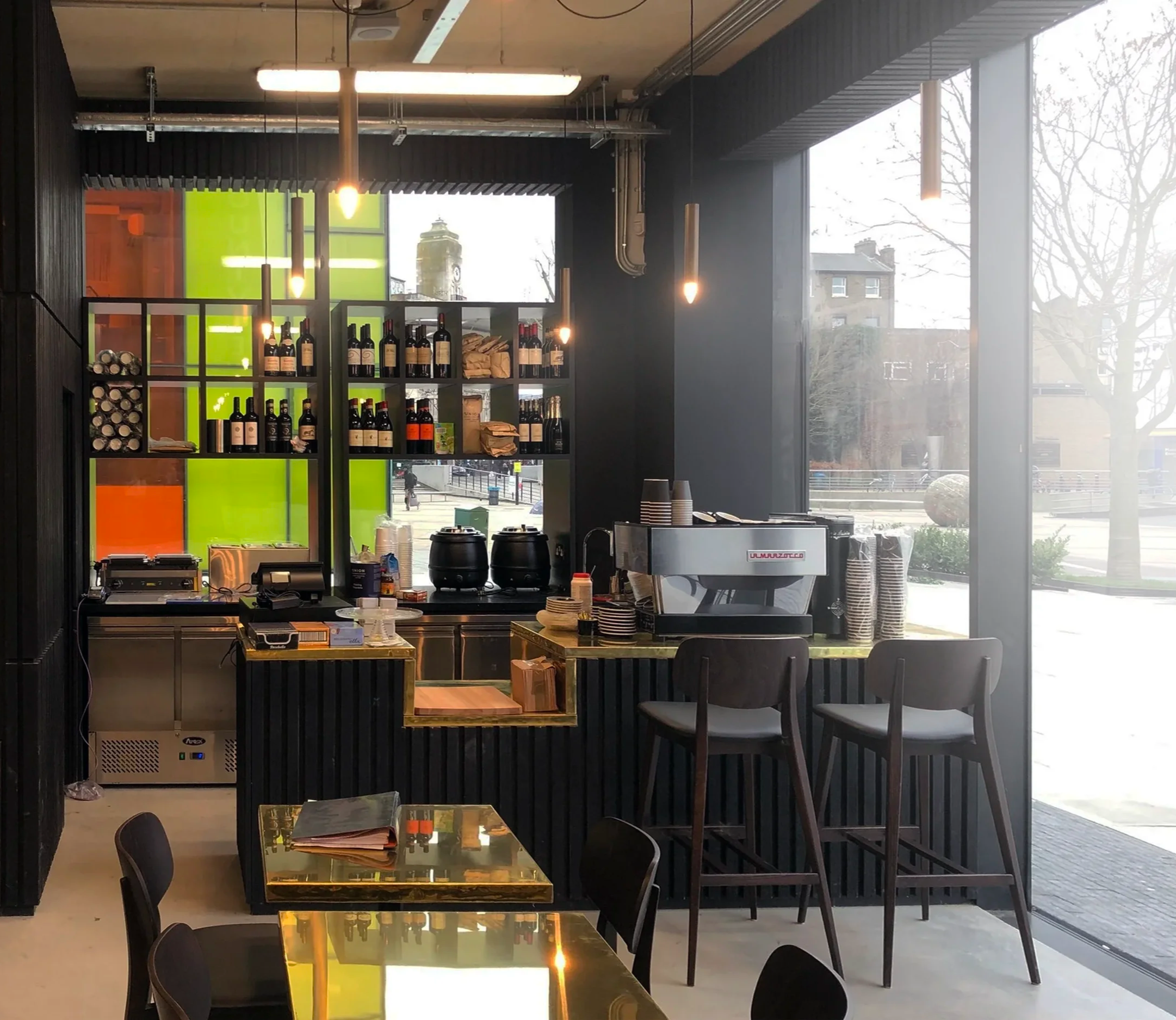 Inside a cafe with black walls, a counter with coffee equipment, shelves with wine bottles and snacks, and a large window with a view of outdoor trees and buildings.