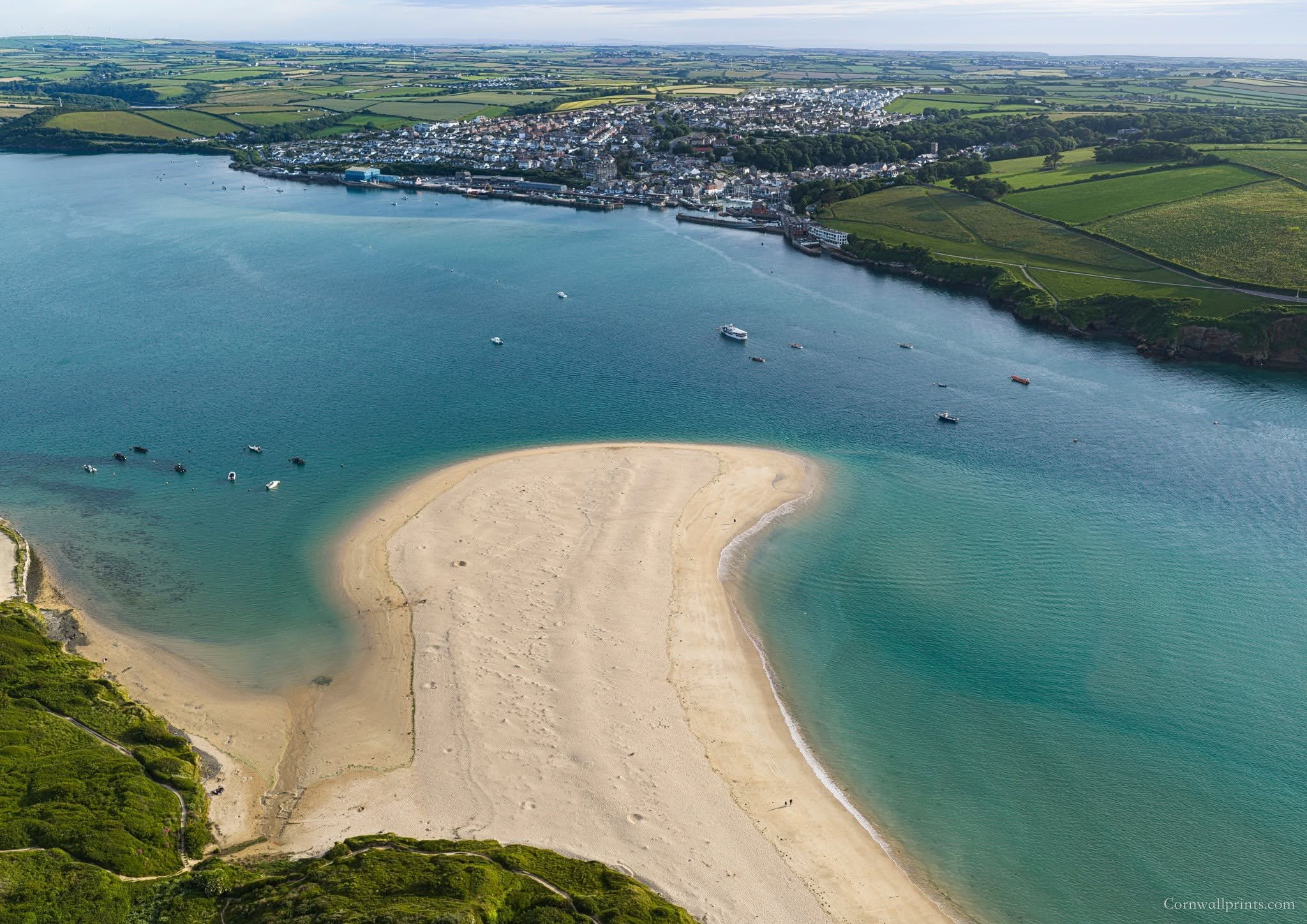 An aerial view of a coastal town with a sandy beach, boats in the water, and green fields surrounding the area.