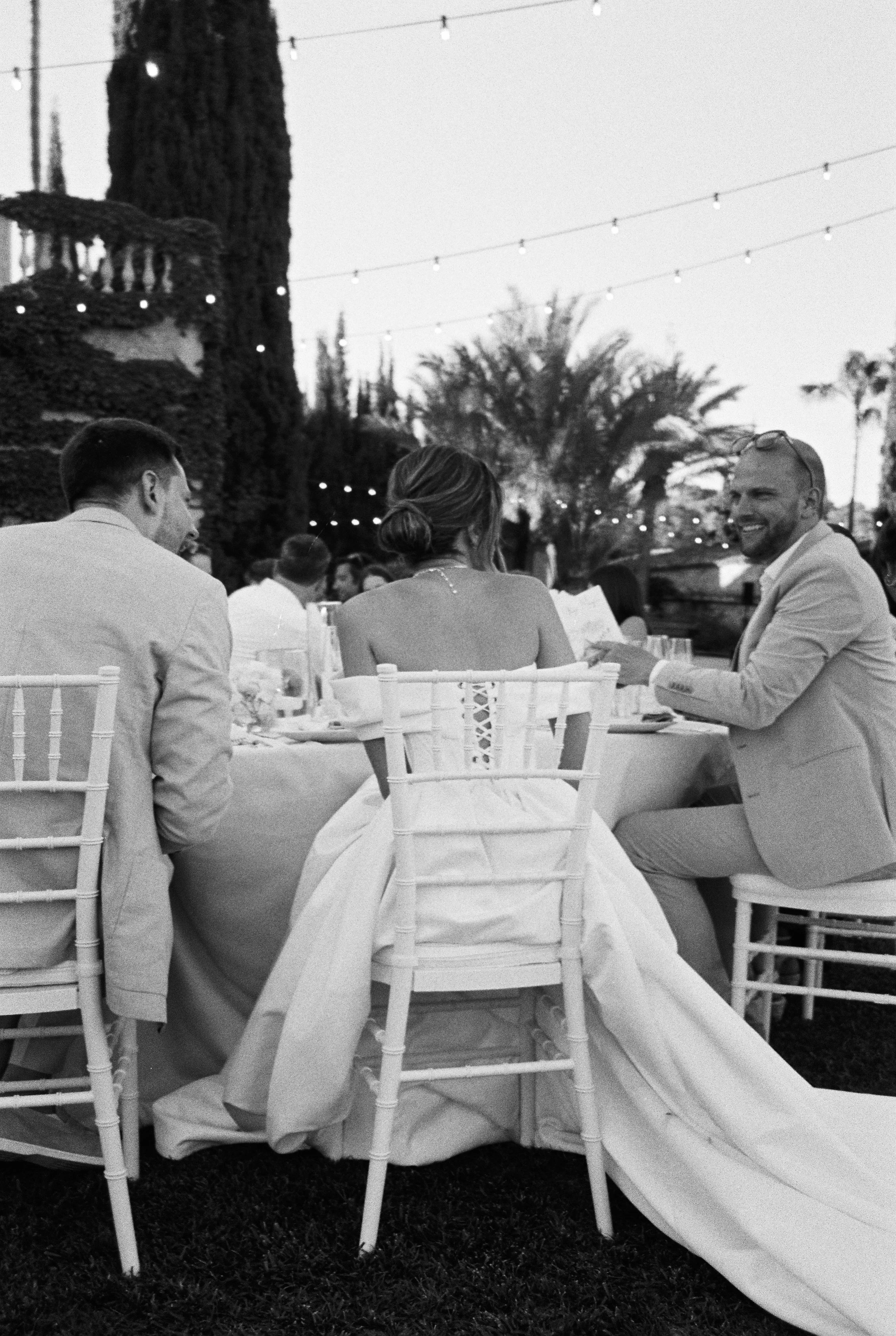Black and white photo of a wedding reception outdoors at dusk with string lights overhead, featuring a bride in a strapless gown and two grooms in suits seated at a table with other guests, palm trees in the background.