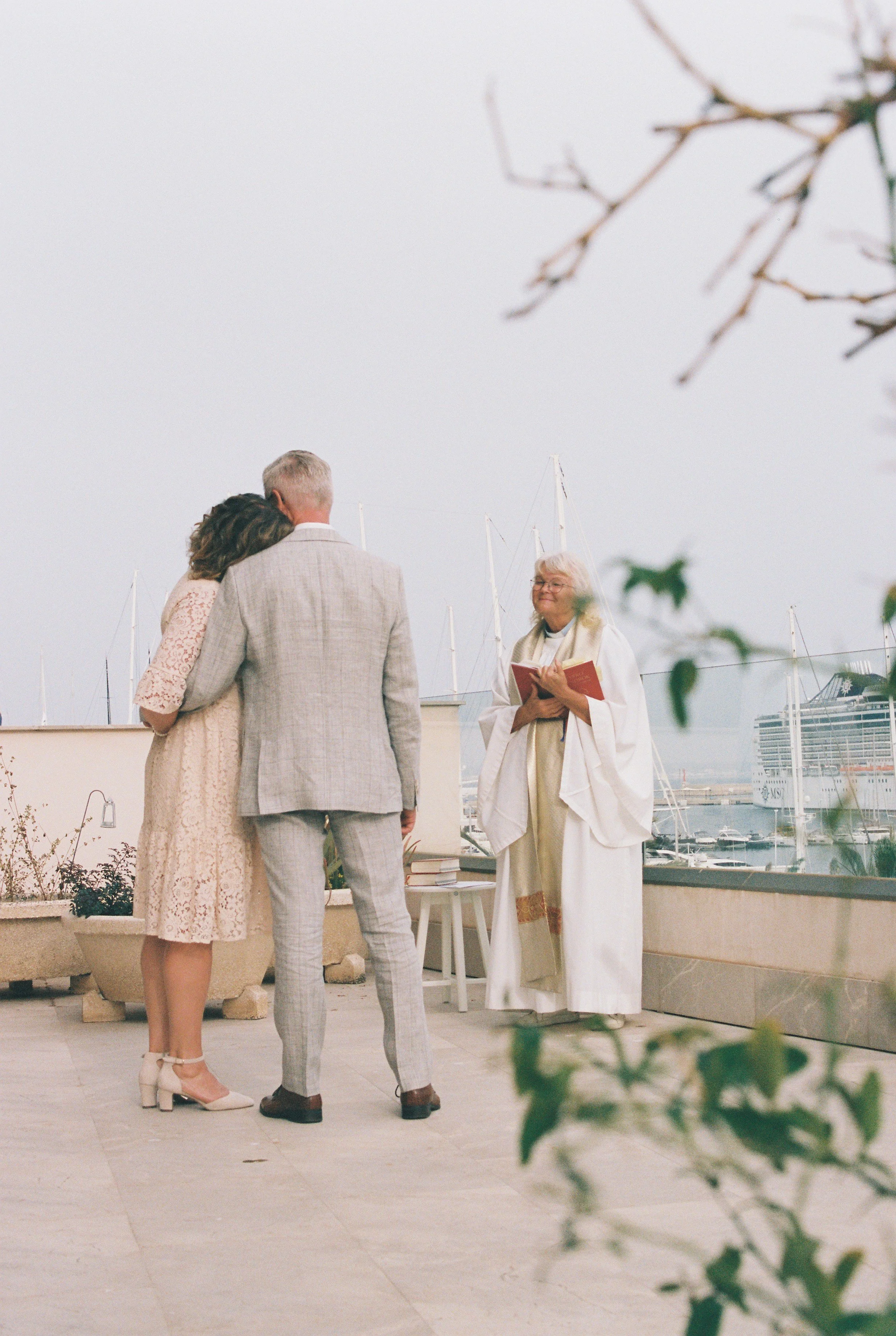 A couple standing with their heads resting against each other during a wedding ceremony outdoors near a marina, with a woman officiant reading from a book. Boats and yachts are visible in the background.