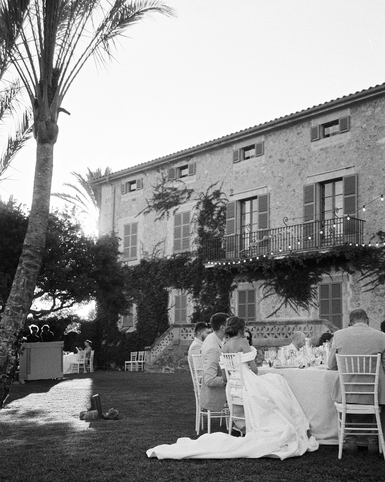 Black and white photo of an outdoor wedding reception at a large stone house with shutters. The bride and groom are seated at a table, surrounded by guests. String lights are visible, and palm trees frame the scene.