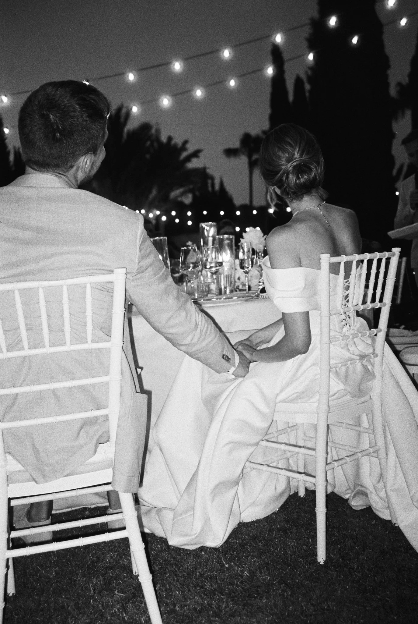 A black-and-white photo of a couple sitting at a banquet table outdoors during evening, with string lights above them. The woman is wearing an off-shoulder dress, and the man is dressed in a light-colored suit. They are holding hands, with the woman’