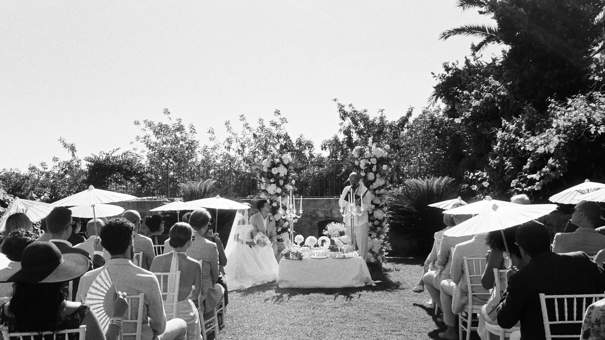 Black and white photo of an outdoor wedding ceremony with guests seated under white umbrellas, facing a decorated altar with flowers and candles, with two people standing at the altar, trees, and bushes in the background.