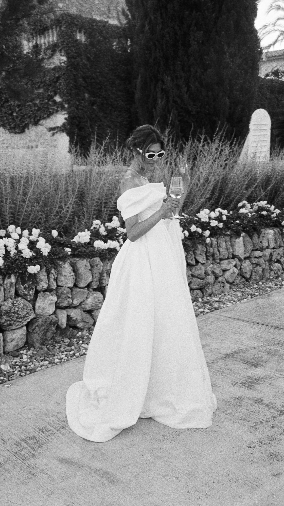 Woman in a white, off-shoulder gown wearing sunglasses, holding a glass of wine, standing outdoors beside a stone flower bed and garden plants.