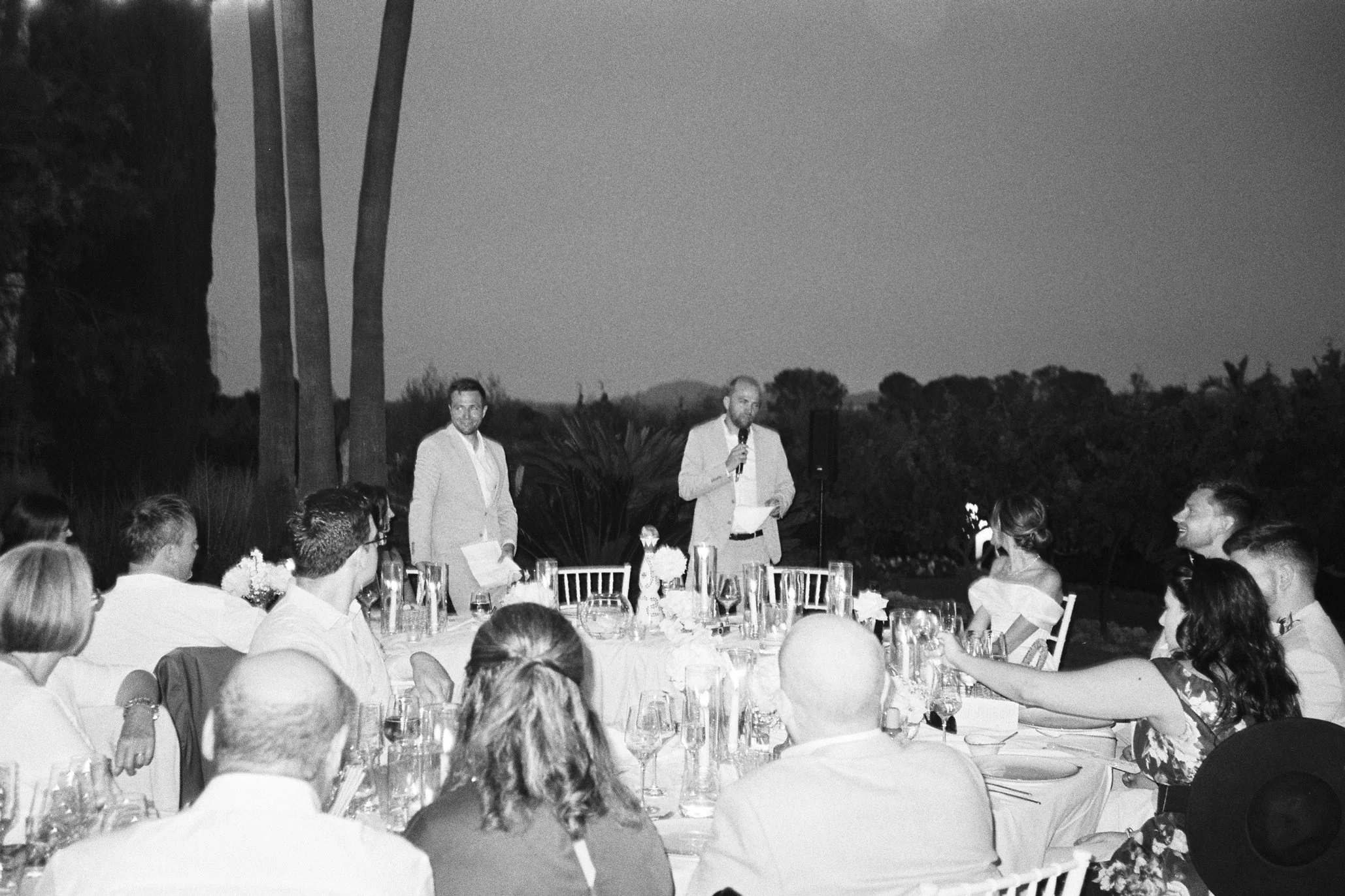 A black-and-white photo of a wedding reception outdoors during the evening. Guests sit around a table with floral centerpieces and glassware. One man stands and speaks into a microphone, while another stands nearby, both dressed in suits.