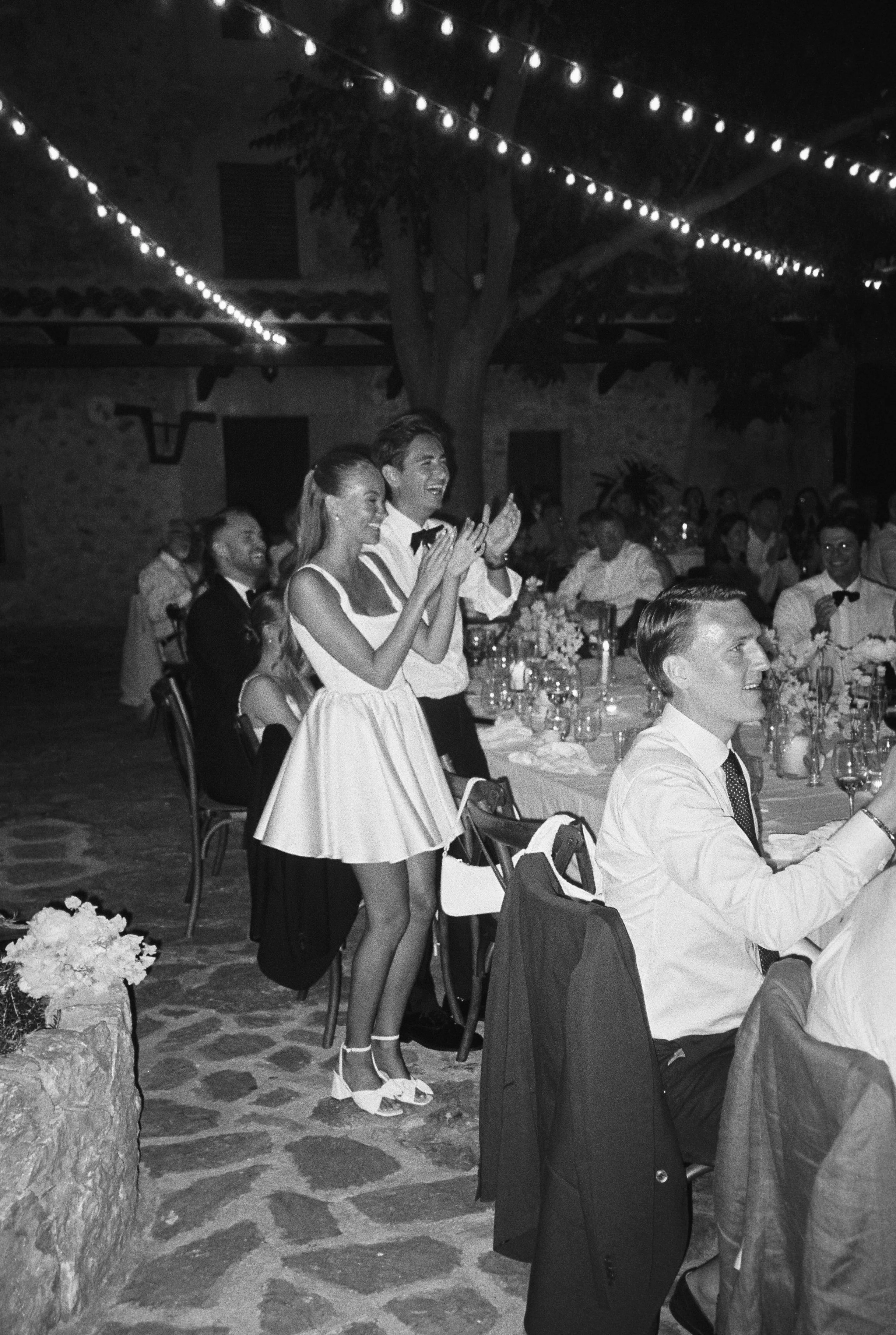 Black and white photo of a lively indoor wedding reception with guests seated at decorated tables, showing a young woman in a white dress and a man in a shirt and tie clapping and smiling, while others in the background also enjoy the celebration wit