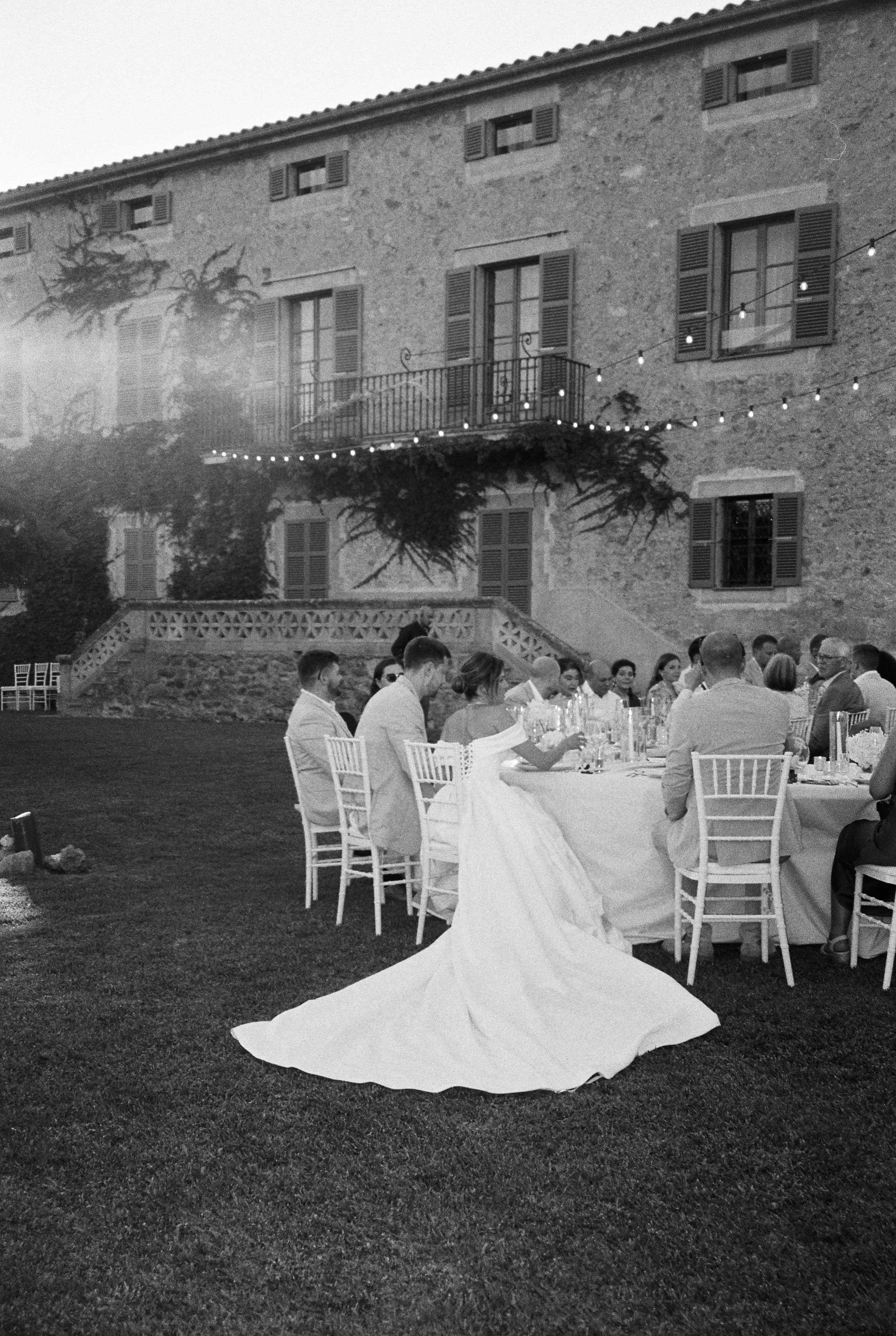 Black and white photo of a wedding reception outdoors in front of a rustic building with string lights and children sitting at a long table. The bride in a wedding gown with a long train is seated at the table.