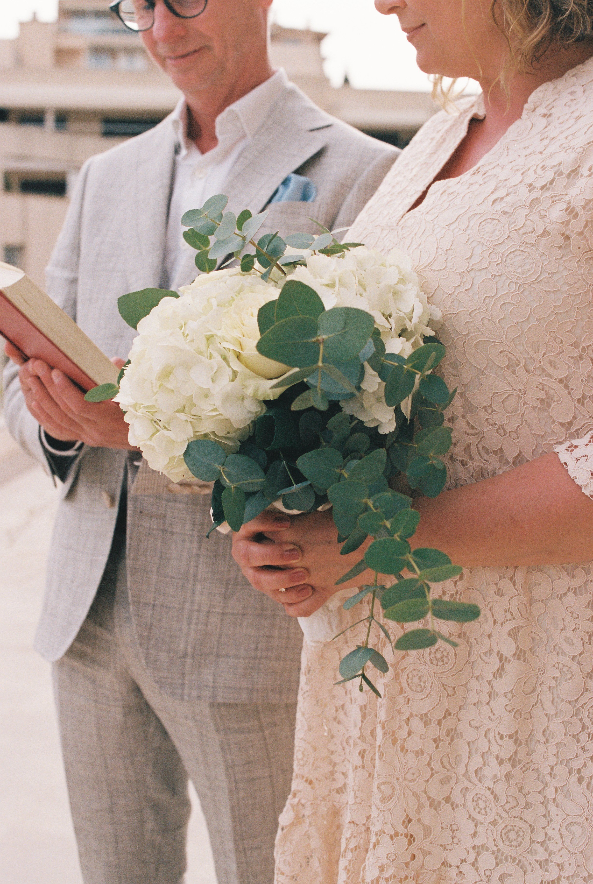 A woman holding a bouquet of white flowers and greenery during a wedding ceremony, with a man reading from a book beside her.