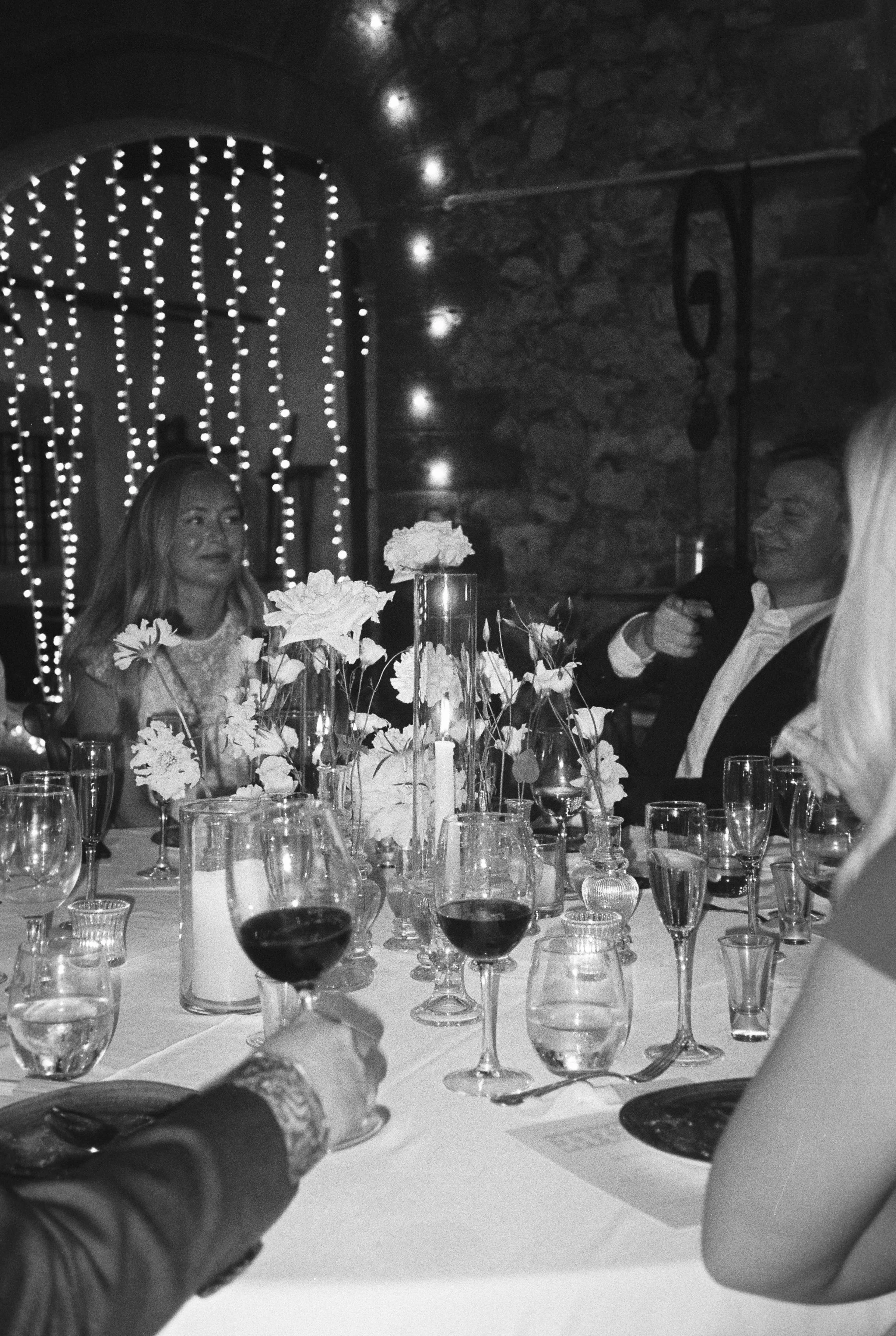 People sitting at a table during a celebration or dinner, with wine glasses, flowers, and candles, in a dimly lit room with string lights and a fireplace.