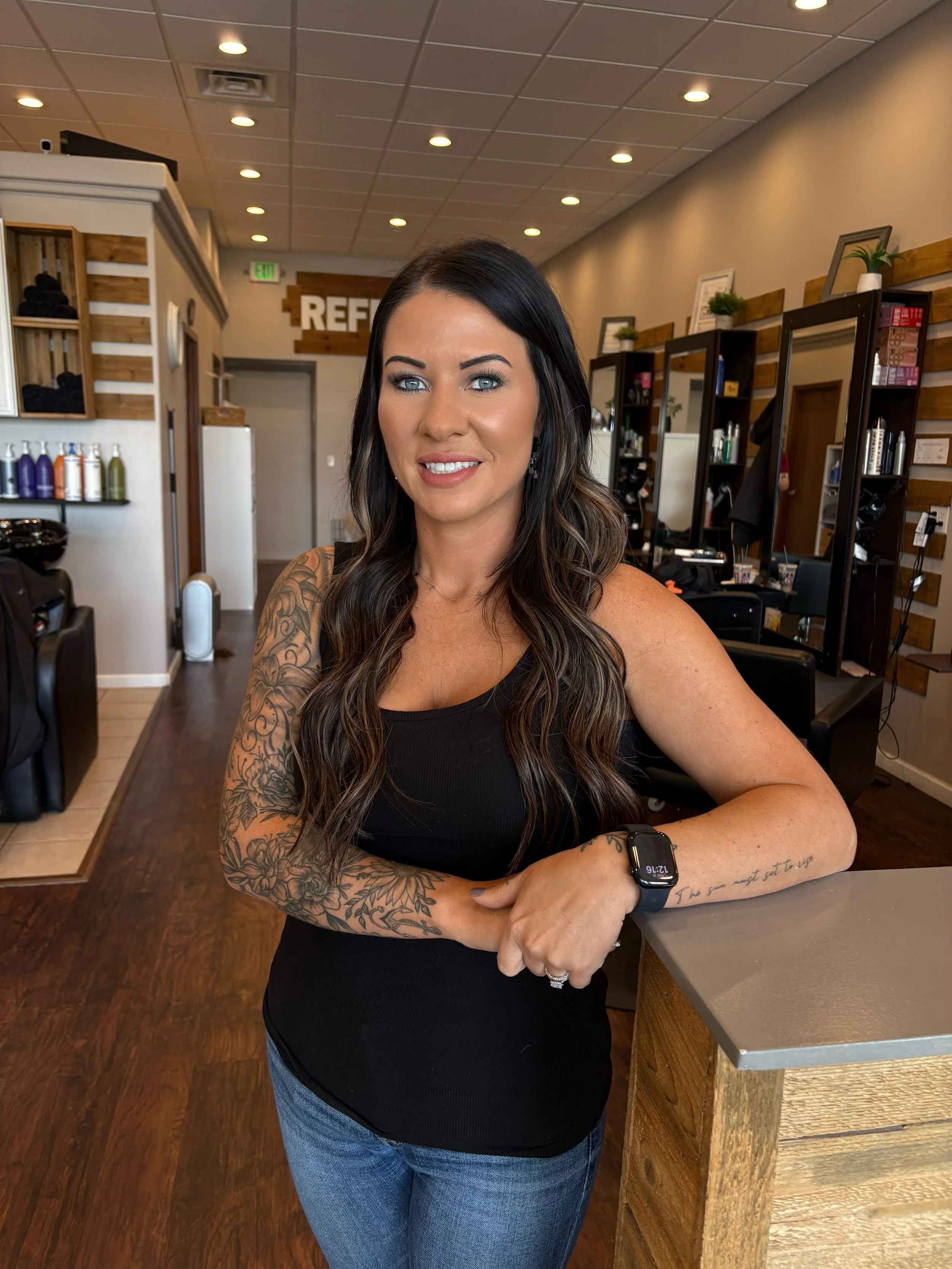 A woman with long dark wavy hair, tattoos on her arms, wearing a black sleeveless top, blue jeans, and a smartwatch, standing inside a modern hair salon with wooden and white decor, mirrors, and hair products.