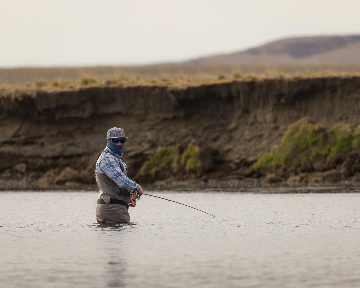 En person fisker i en flod eller sø, iført waders, hat, solbriller og ansigtsskærm, omgivet af et naturlandskab med bakker og klipper.