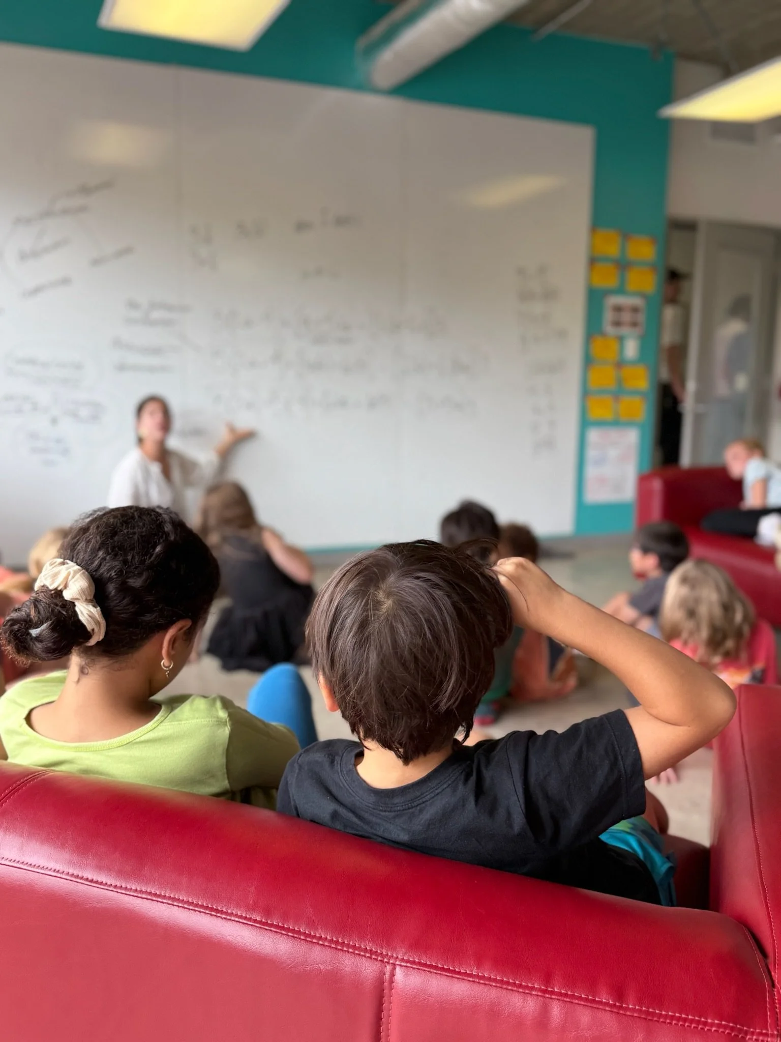A teacher is in the background kneeling on the ground and pointing to math expressions on a board. Many students sit in front of her actively listening.