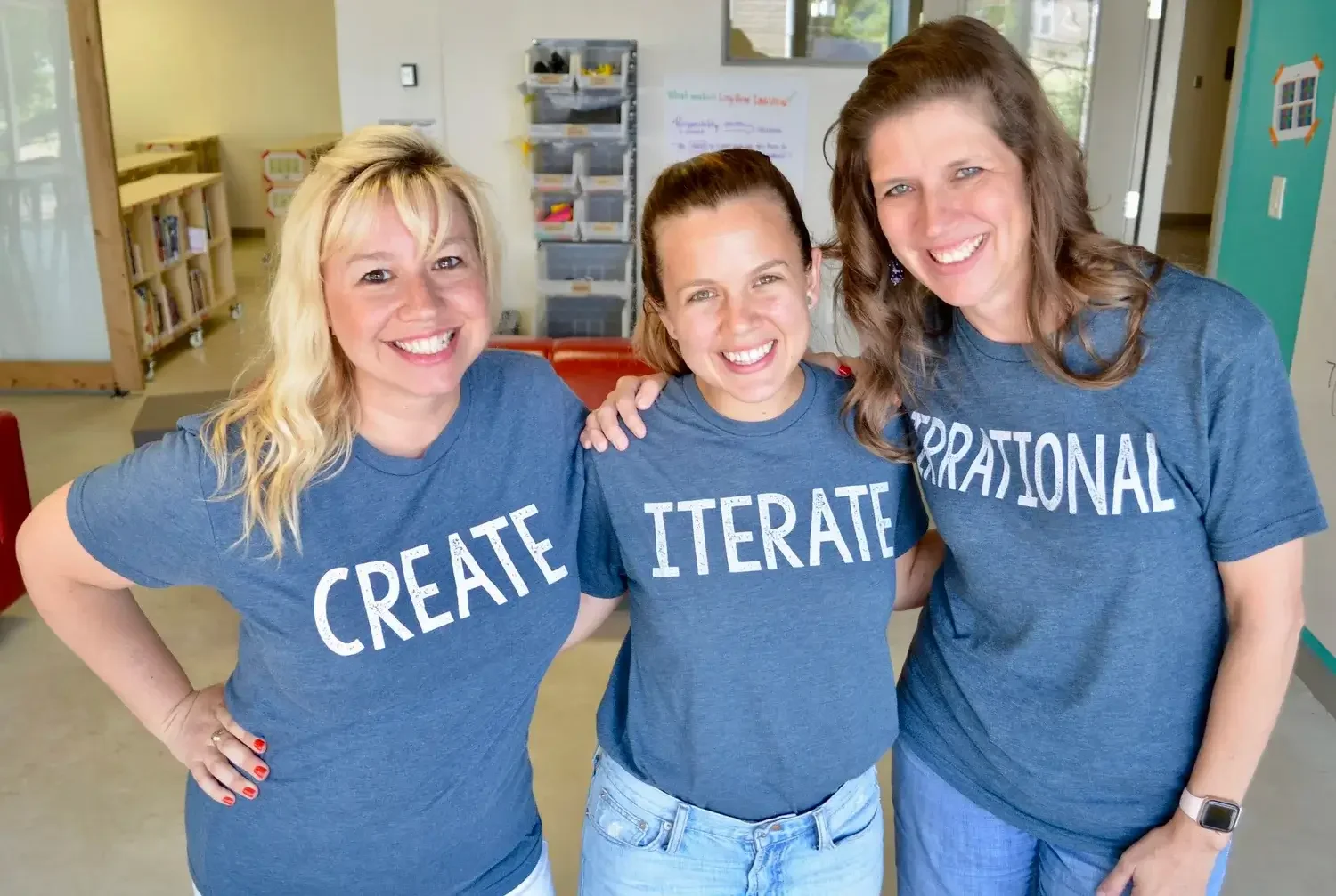 Three women standing together in a classroom, all wearing matching blue T-shirts with the word 'CREATE' printed on them, smiling and embracing each other.