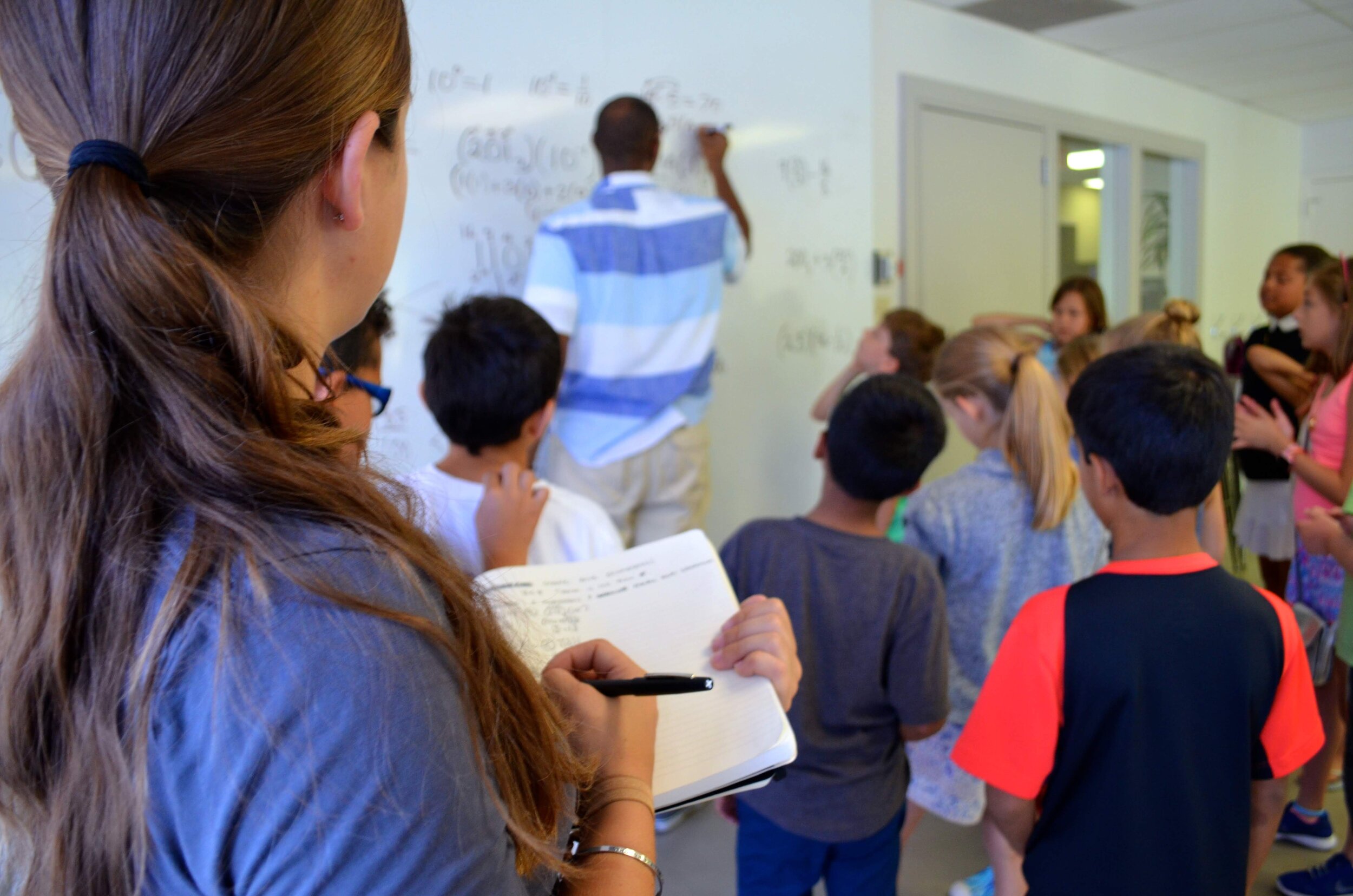 A teacher writes on a whiteboard while a group of children observe. A coach makes notes.