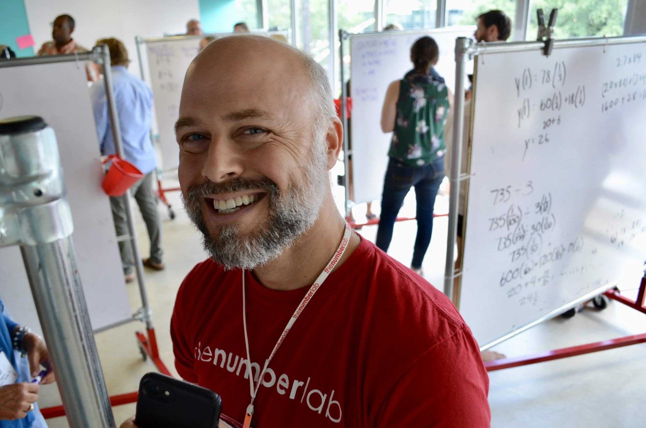 Man faces camera and smiles. He is wearing a red shirt and has a beard. There are people behind him at whiteboards.