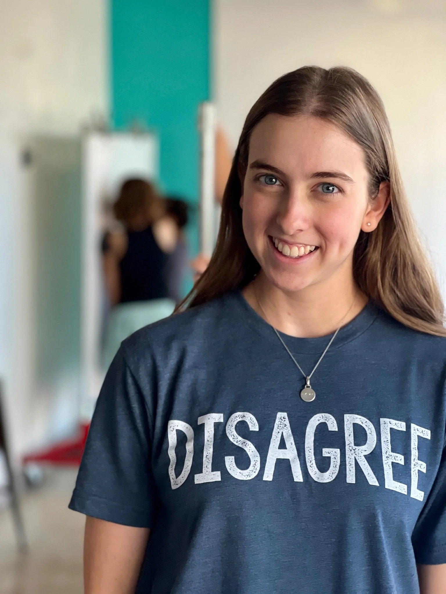 A woman in a blue shirt that says 'disagree' faces the camera and smiles. In the background are people at whiteboards.