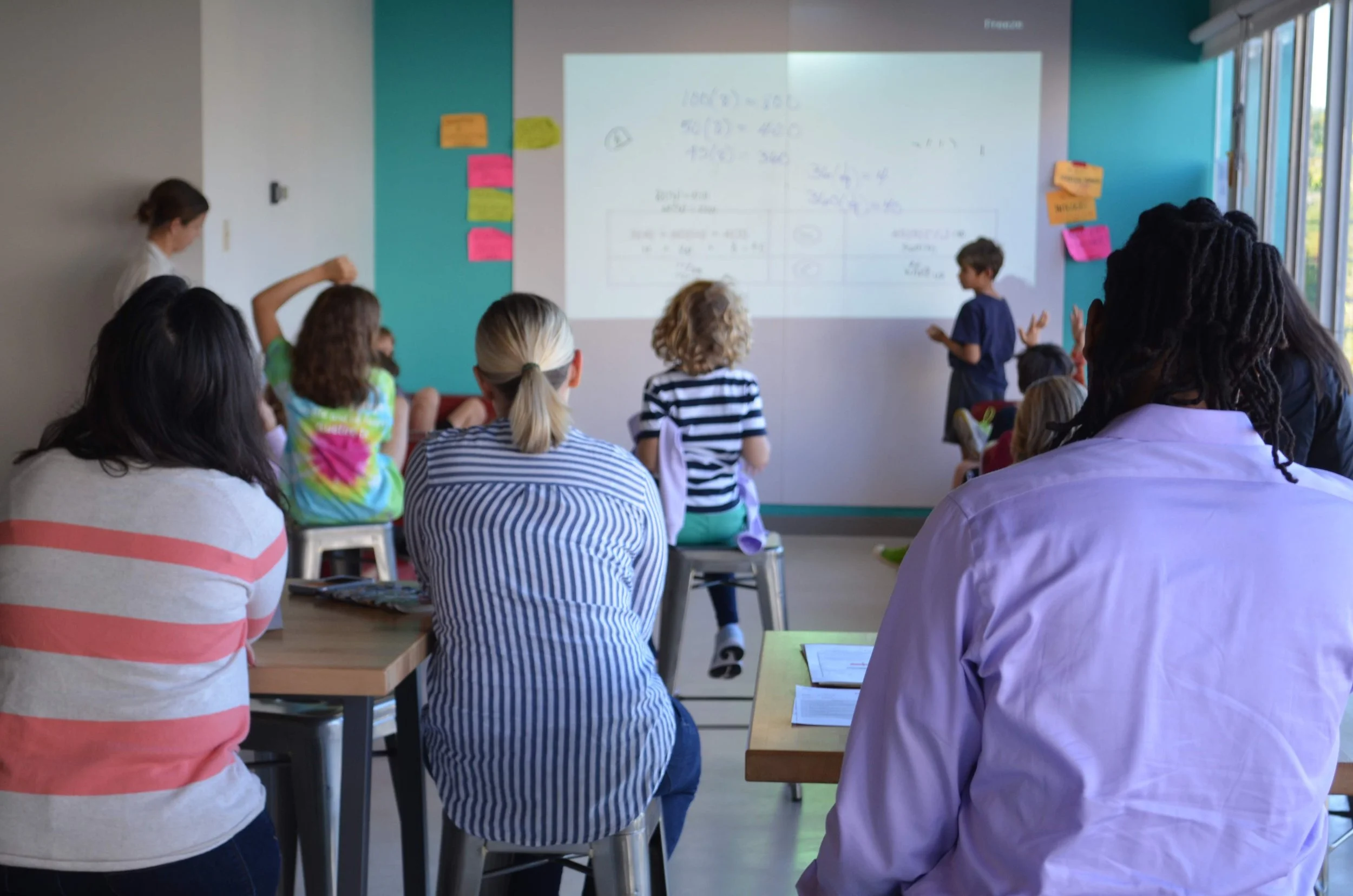A group of math teachers observe a lesson. Children are at the whiteboard talking and discussing.