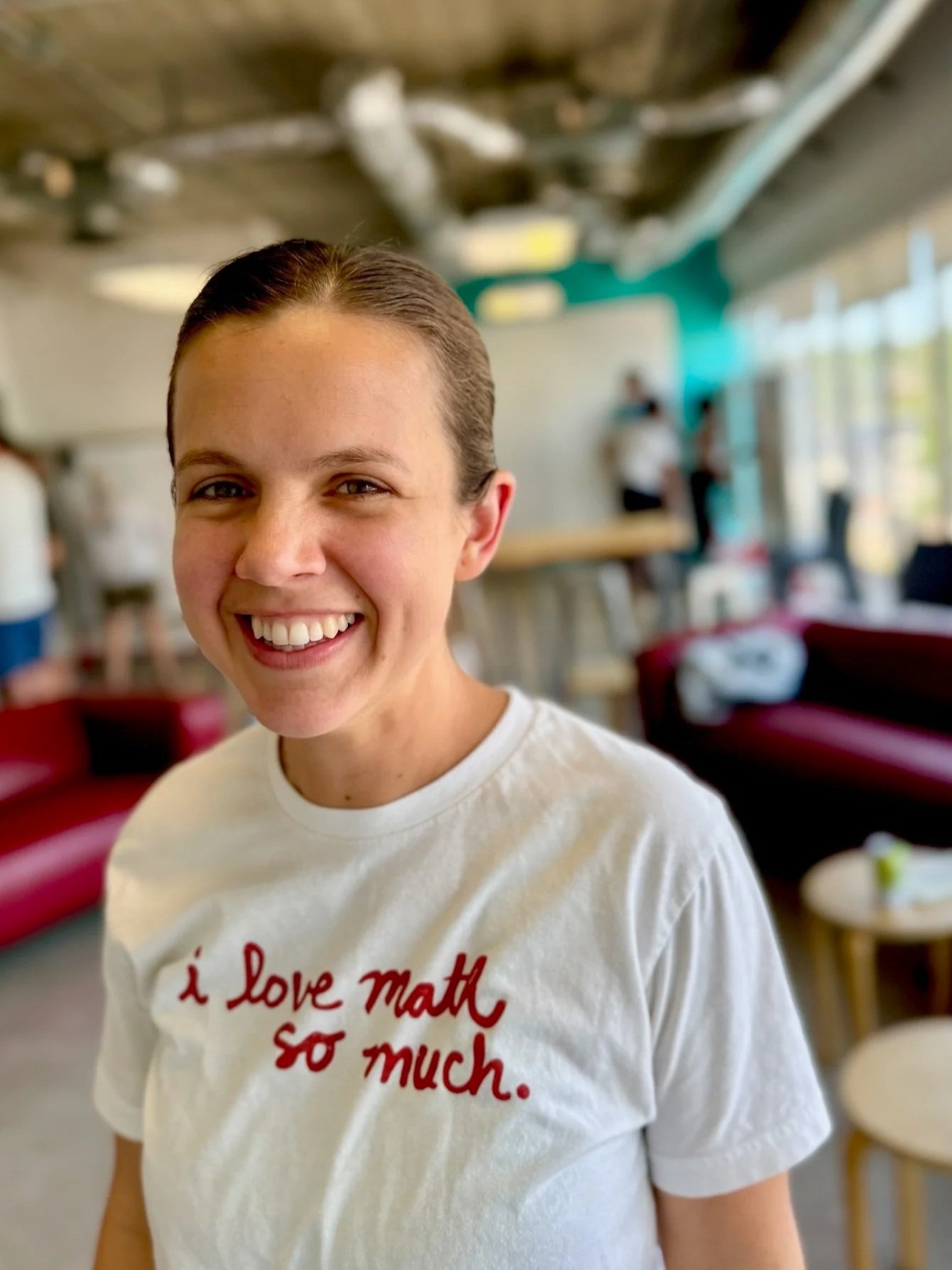 Woman in white shirt looks at camera and smiles. Behind her is a busy room of people, red sofas, stools, and whiteboards.