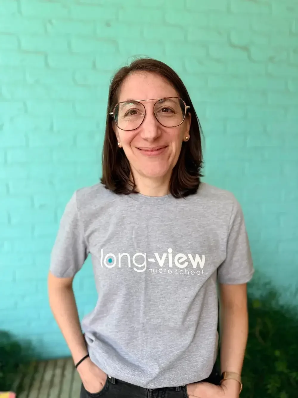 Woman stands in front of turquoise wall and smiles at the camera. She is wearing a t-shirt that says "Long-View Micro School."
