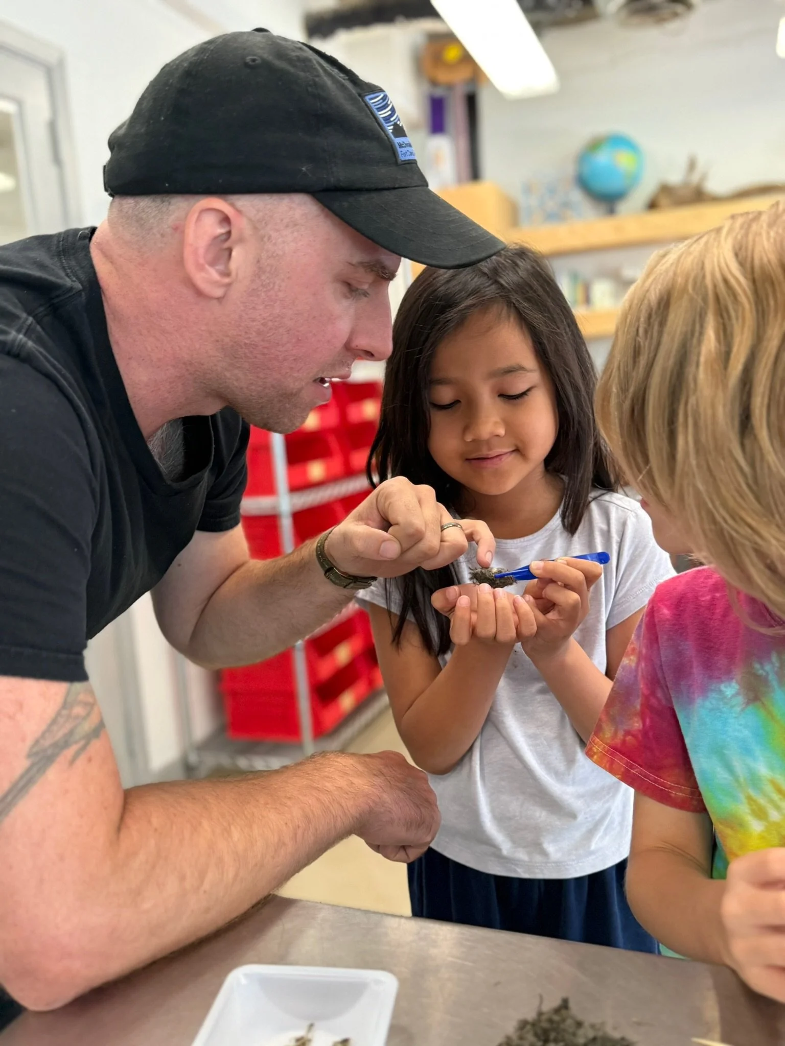 A man in a black shirt and black baseball camp is talking to two children. The child in a white shirt is holding something and the man is looking carefully and pointing. Behind them are red bins and classroom items.