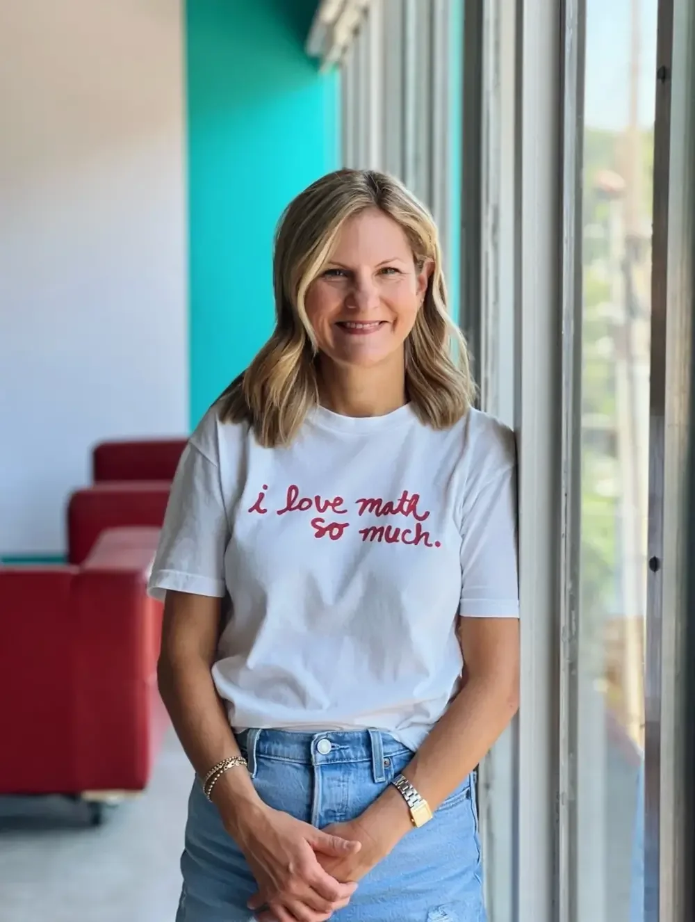 Woman in white shirt that says 'I love math so much' leans against large window and smiles. In the background are red sofas, whiteboard, and turquoise wall.