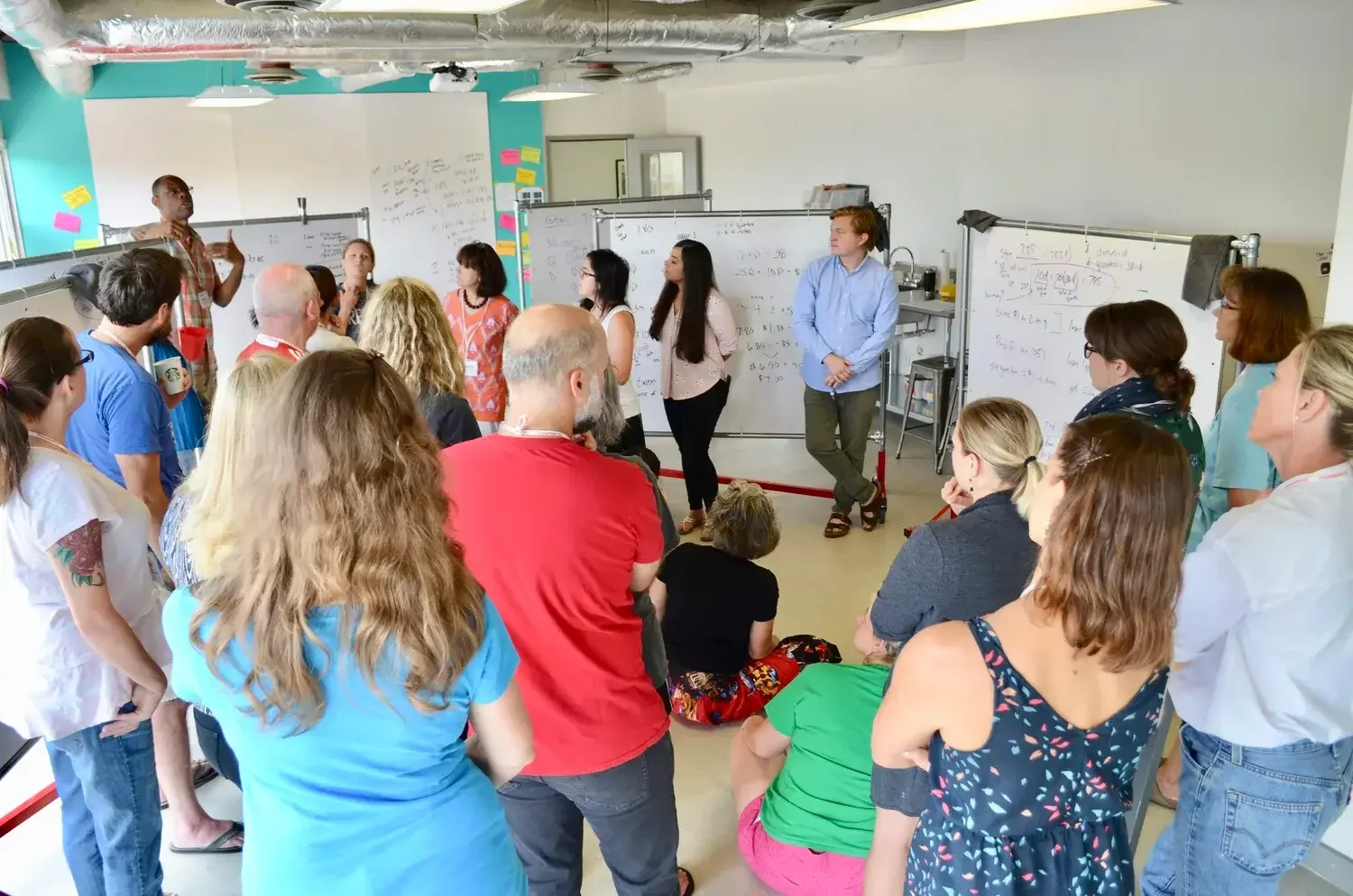 A large group of math teachers gathers in a room of whiteboards. A consultant speaks about math.