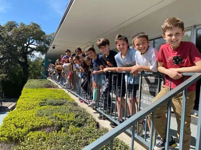 Children line an outdoor porch, standing against a rail. They are all smiling. Sun is shining and green bushes are below them.