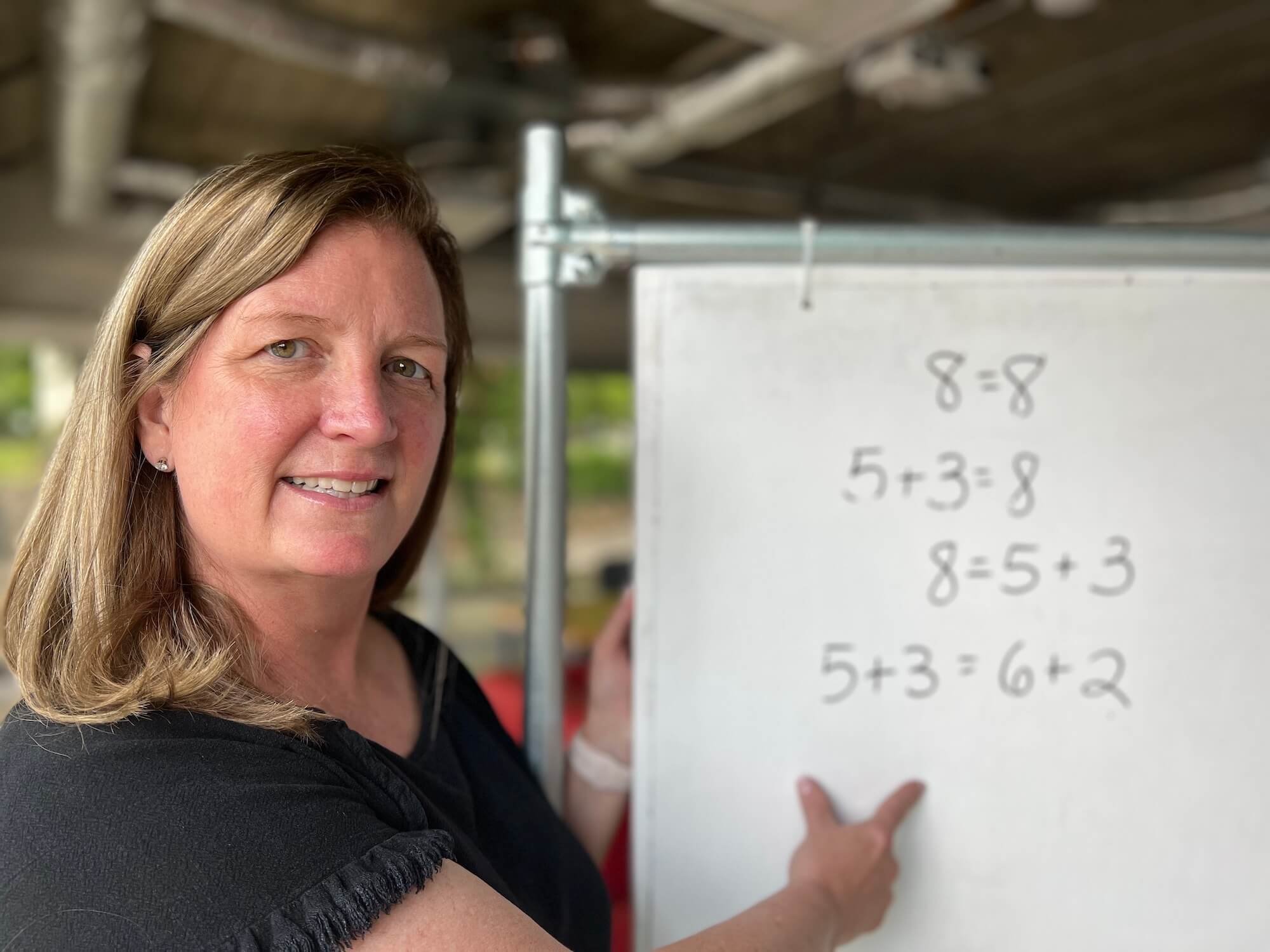 Woman in black shirt faces camera and smiles. She is in an industrial-style room with a window and whiteboard.