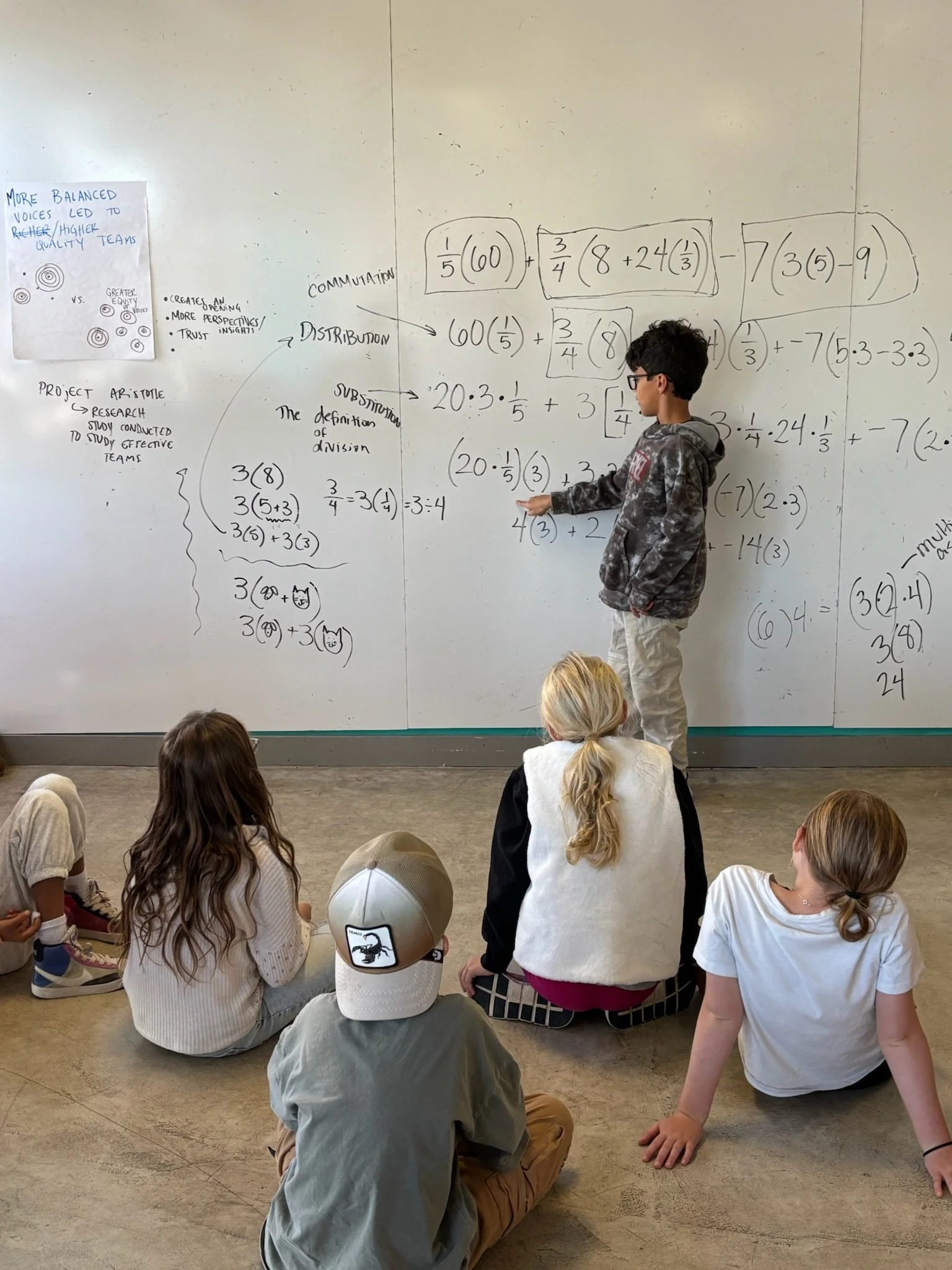 A whiteboard shows a complex mathematical algorithm. A child stands near the whiteboard and points to a term. Other children observe or listen.