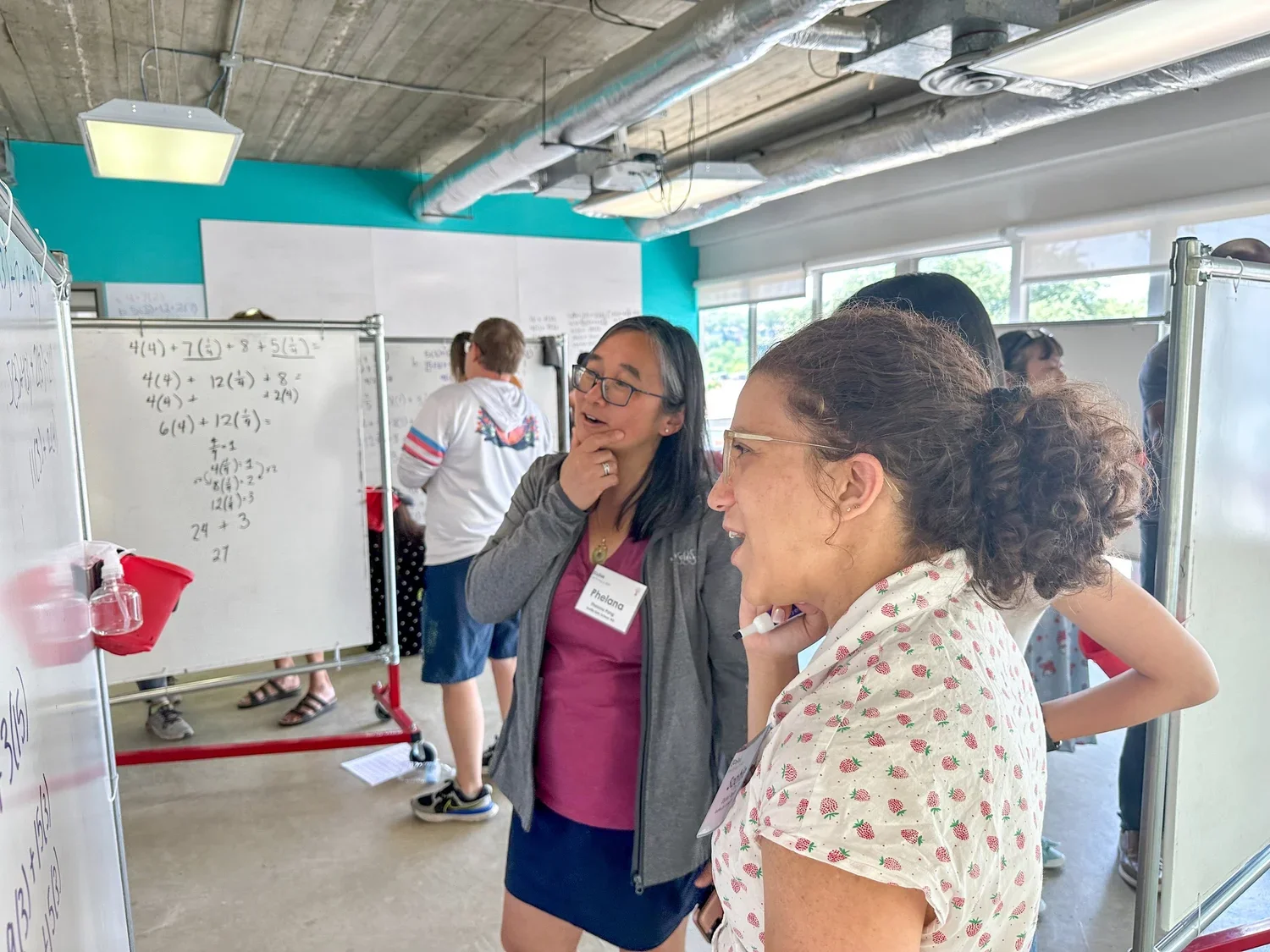 Two women are talking and smiling at an indoor event with whiteboards and other people in the background. One woman has short dark hair and glasses, the other has curly hair and is wearing a strawberry print shirt.
