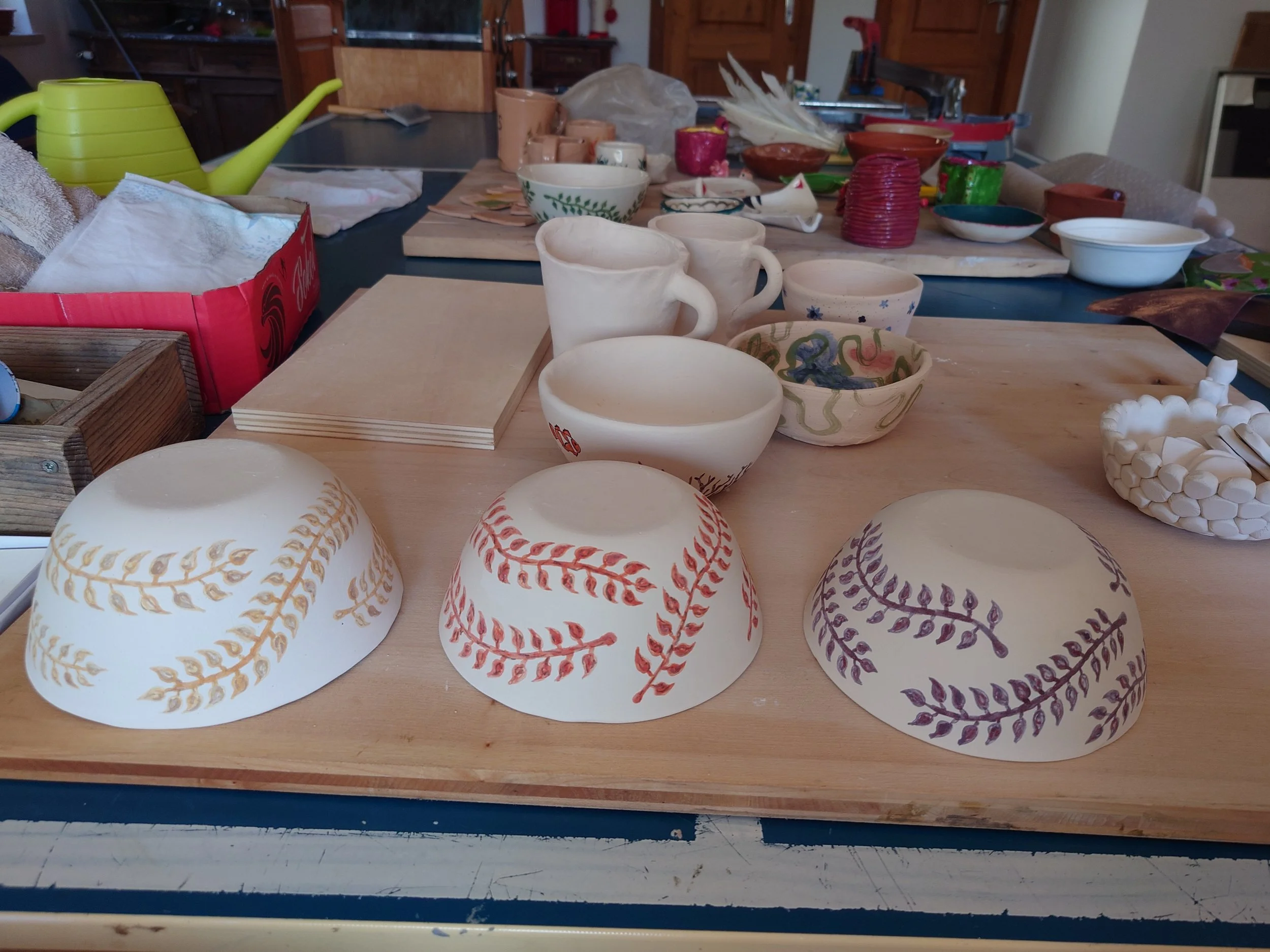A workspace with various handmade pottery pieces, including cups, bowls, and decorative items, some with painted leaf patterns in red, purple, and blue, arranged on a wooden table.