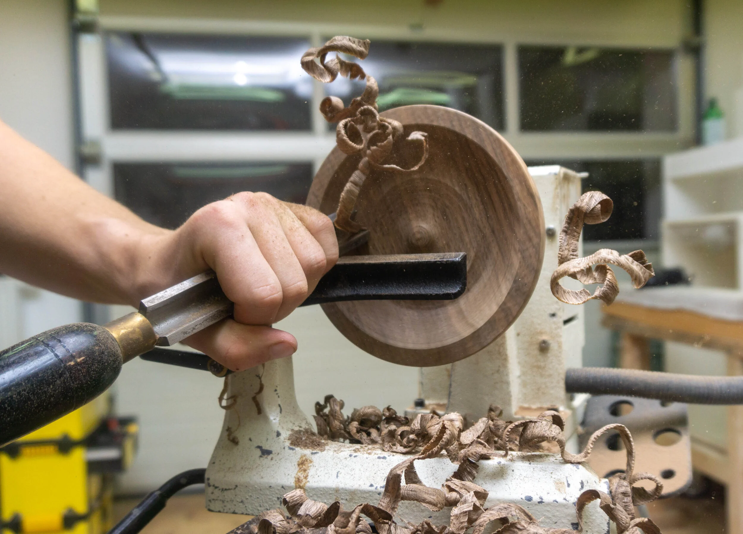 Hands using a woodworking lathe to carve wood shavings from a piece of wood.