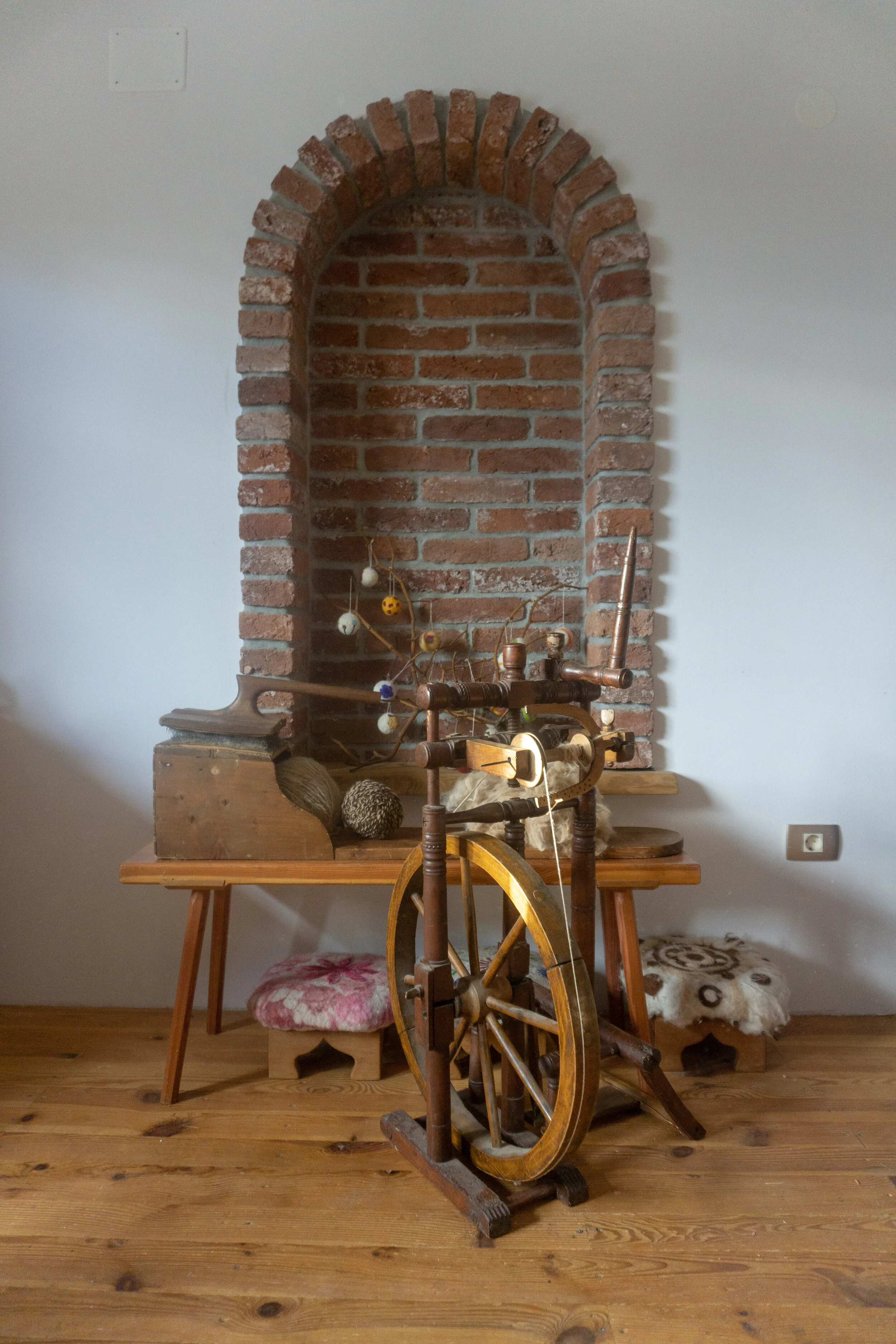 a spinning wheel with some wool, and a "gredeše", tool for combing the wool, in front of exposed red brick wall with arched opening.
