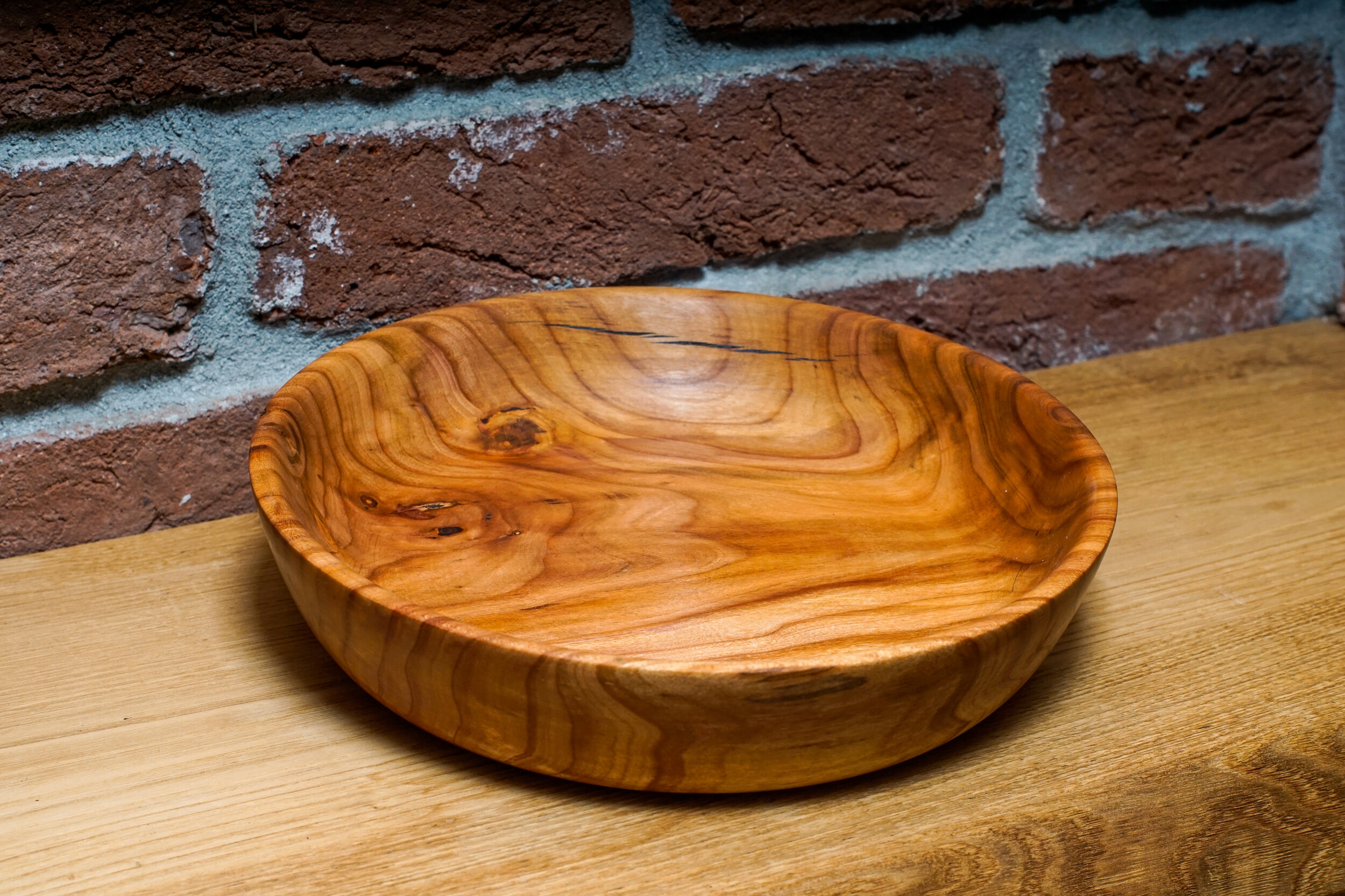 A  turned wooden bowl with a natural wood grain pattern, placed on a wooden surface in front of a red brick wall.
