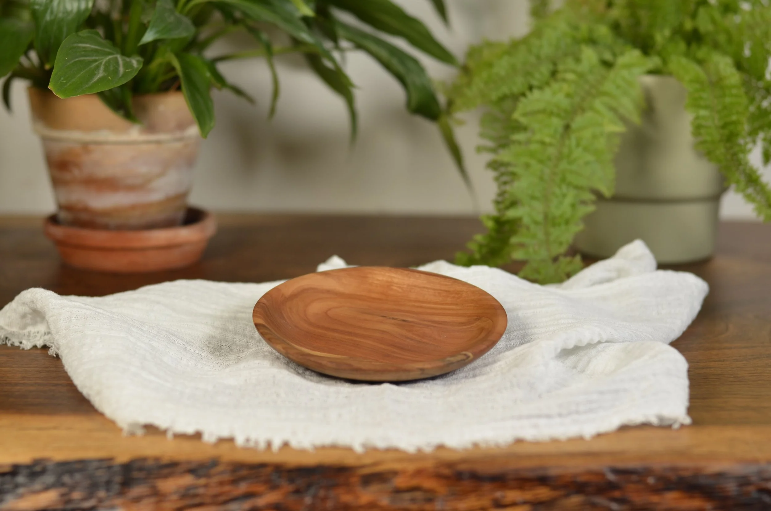 A turned wooden dish placed on a white cloth on a wooden table, with potted green leafy plants in the background.