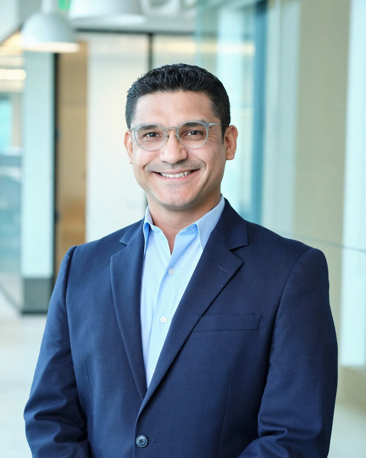 A man in a navy blue suit and glasses smiling in an office setting.