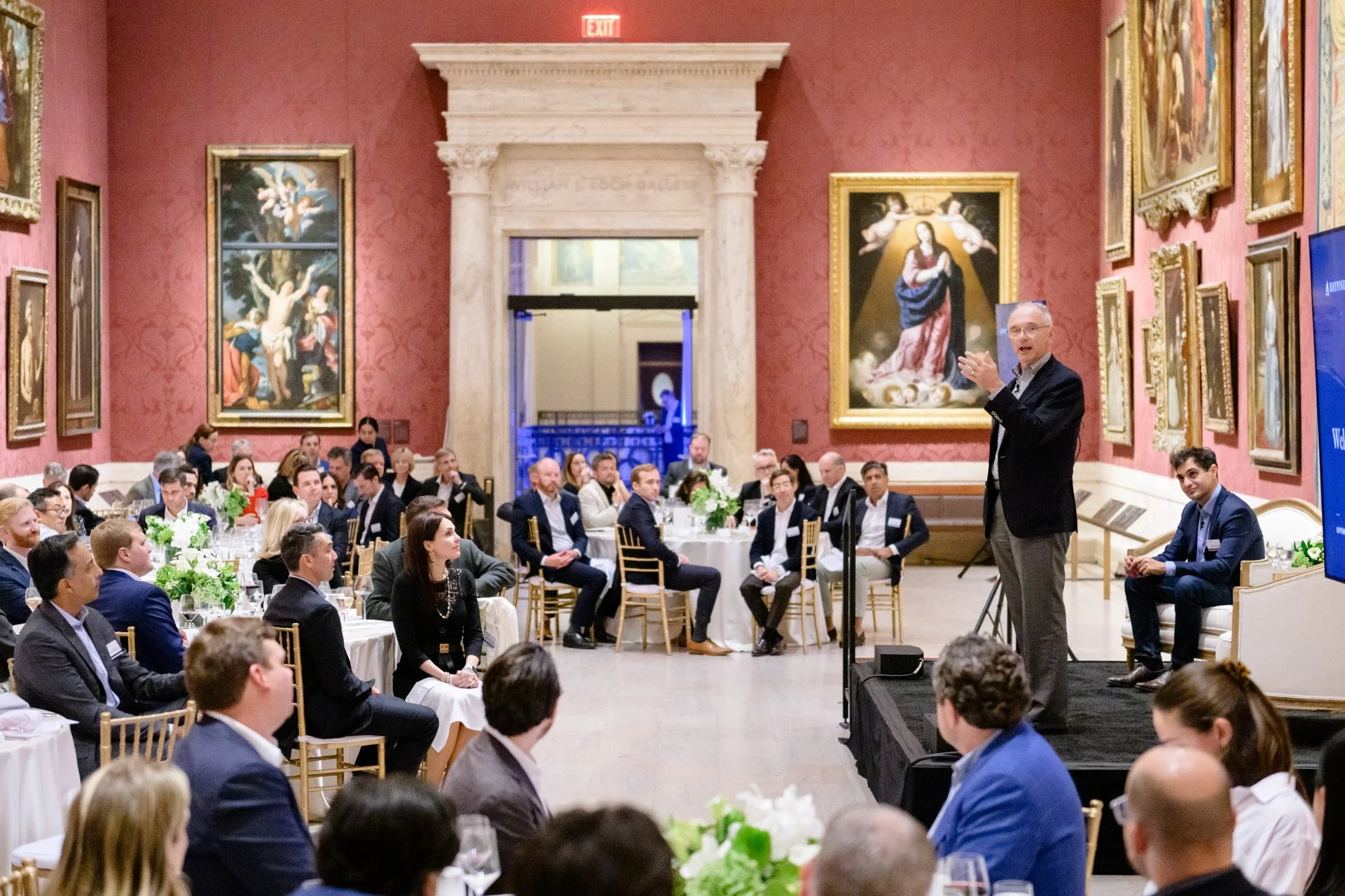 Business conference or formal event in a large, ornate room with red walls decorated with framed religious paintings. A speaker stands on a stage addressing an audience seated at round tables with white tablecloths and floral centerpieces.