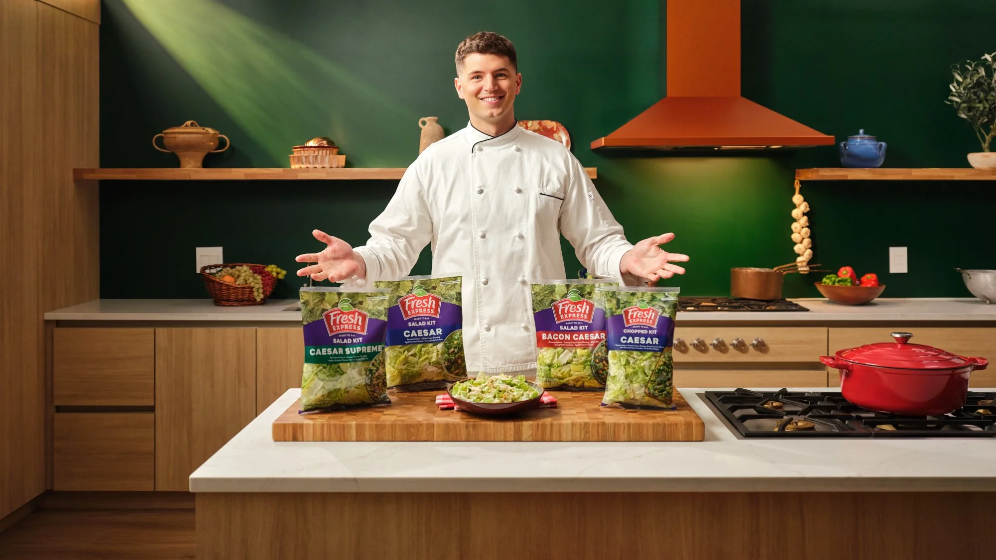 A smiling chef standing in a modern kitchen with wooden cabinets, colorful wall, and a stove, presenting four bags of Caesar salad kits on a wooden cutting board.