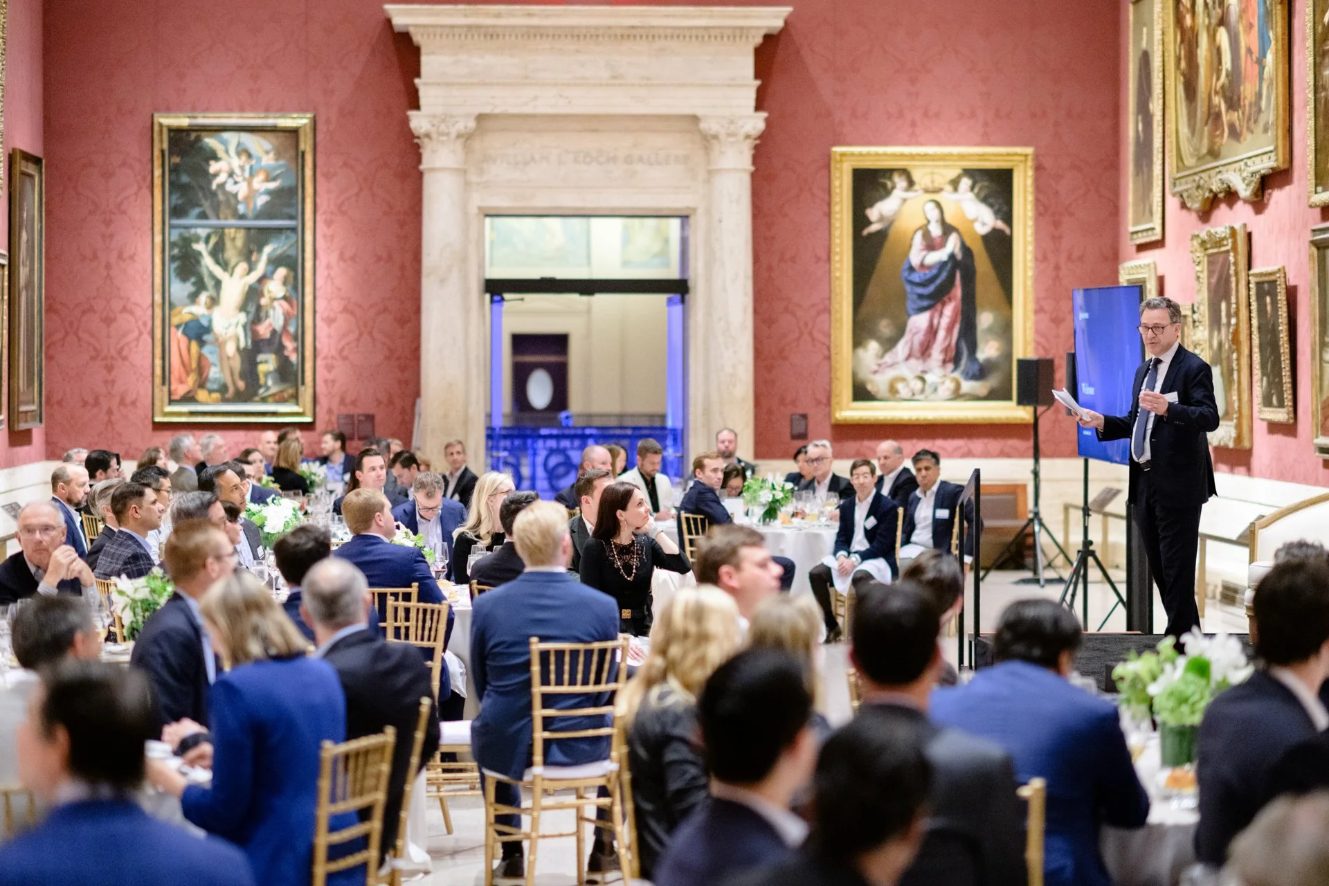 A man in a suit giving a speech at a formal event or conference in a grand room with red walls, ornate paintings, and large columns, with an audience seated at round tables.