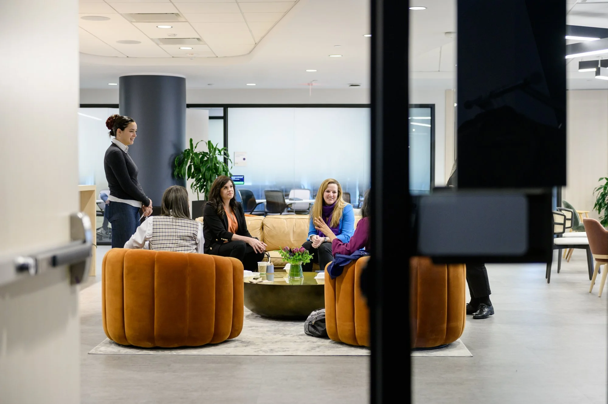 Group of five women socializing in a modern office lounge, seated on orange velvet chairs and a sofa, with coffee cups and a small flower arrangement on the coffee table. One woman stands nearby, smiling.