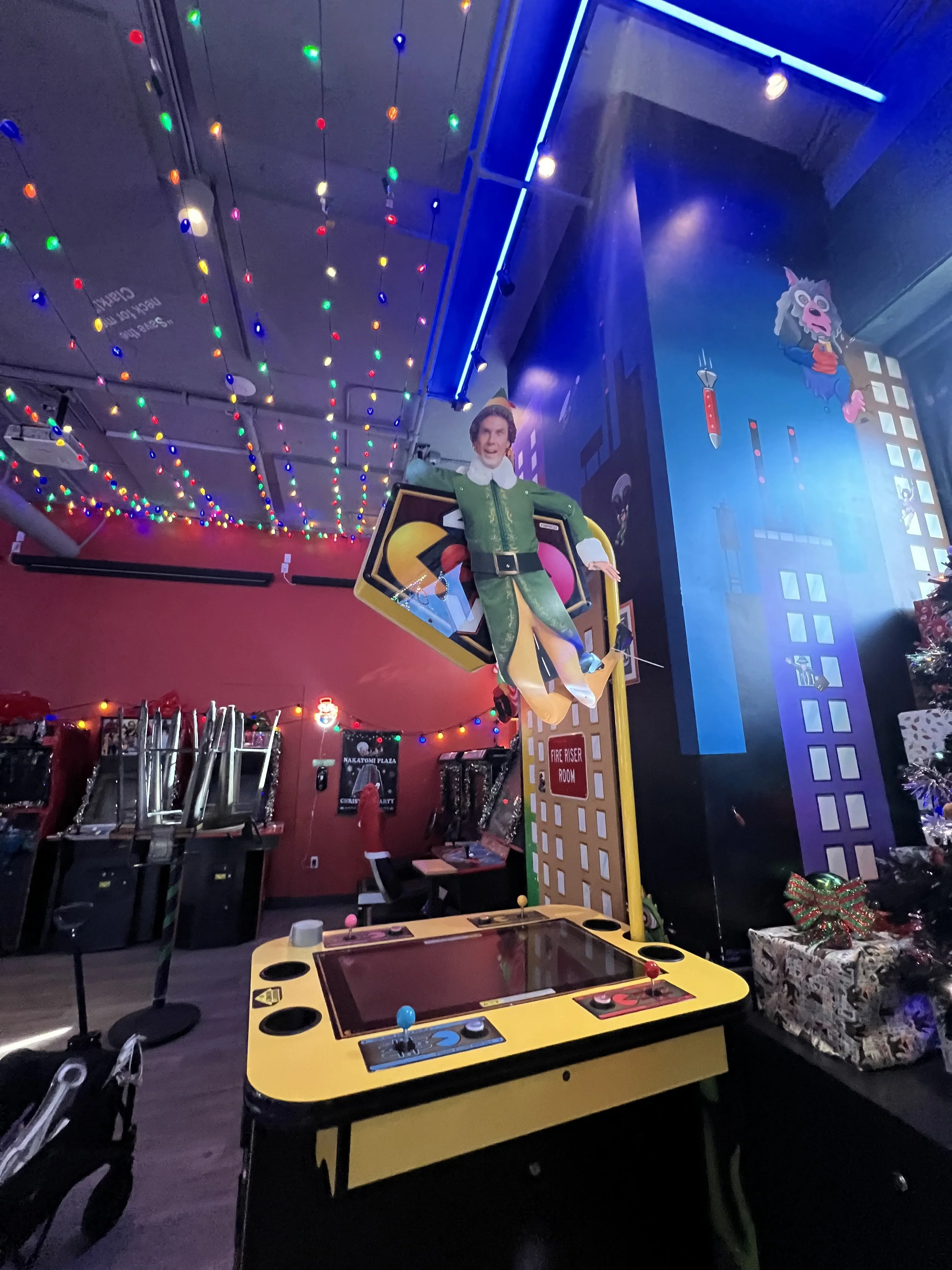 A colorful arcade game with a figure dressed as Buddy the Elf from the movie 'Elf' hanging from a structure. The room is decorated with string lights and a Christmas tree with gifts, creating a festive atmosphere. Mini Boss San Jose. 