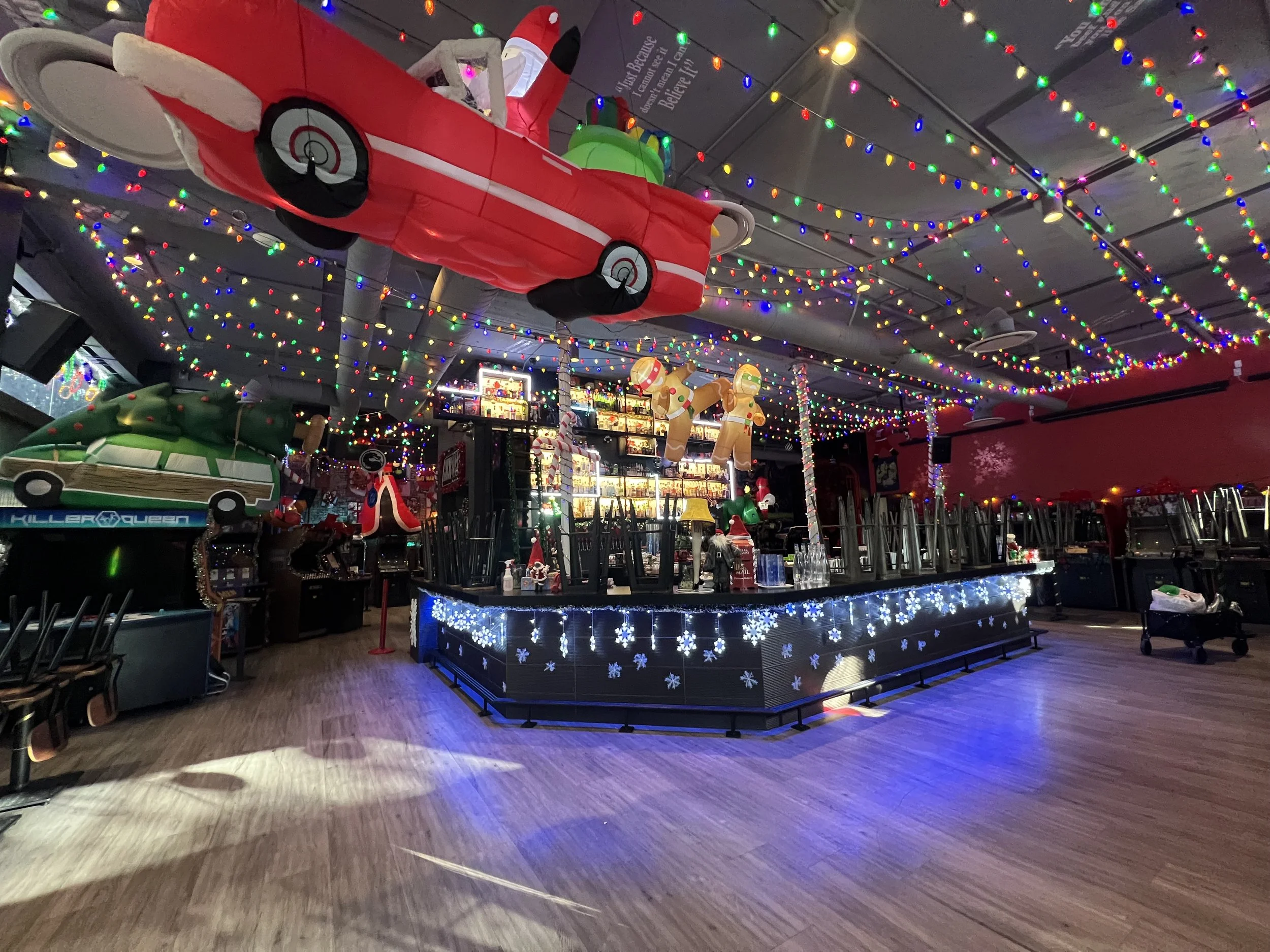 Indoor space decorated for Christmas with colorful string lights, inflatable Christmas objects including a red airplane and gingerbread figures, and a bar area with snowflake designs. Mini Boss San Jose. 
