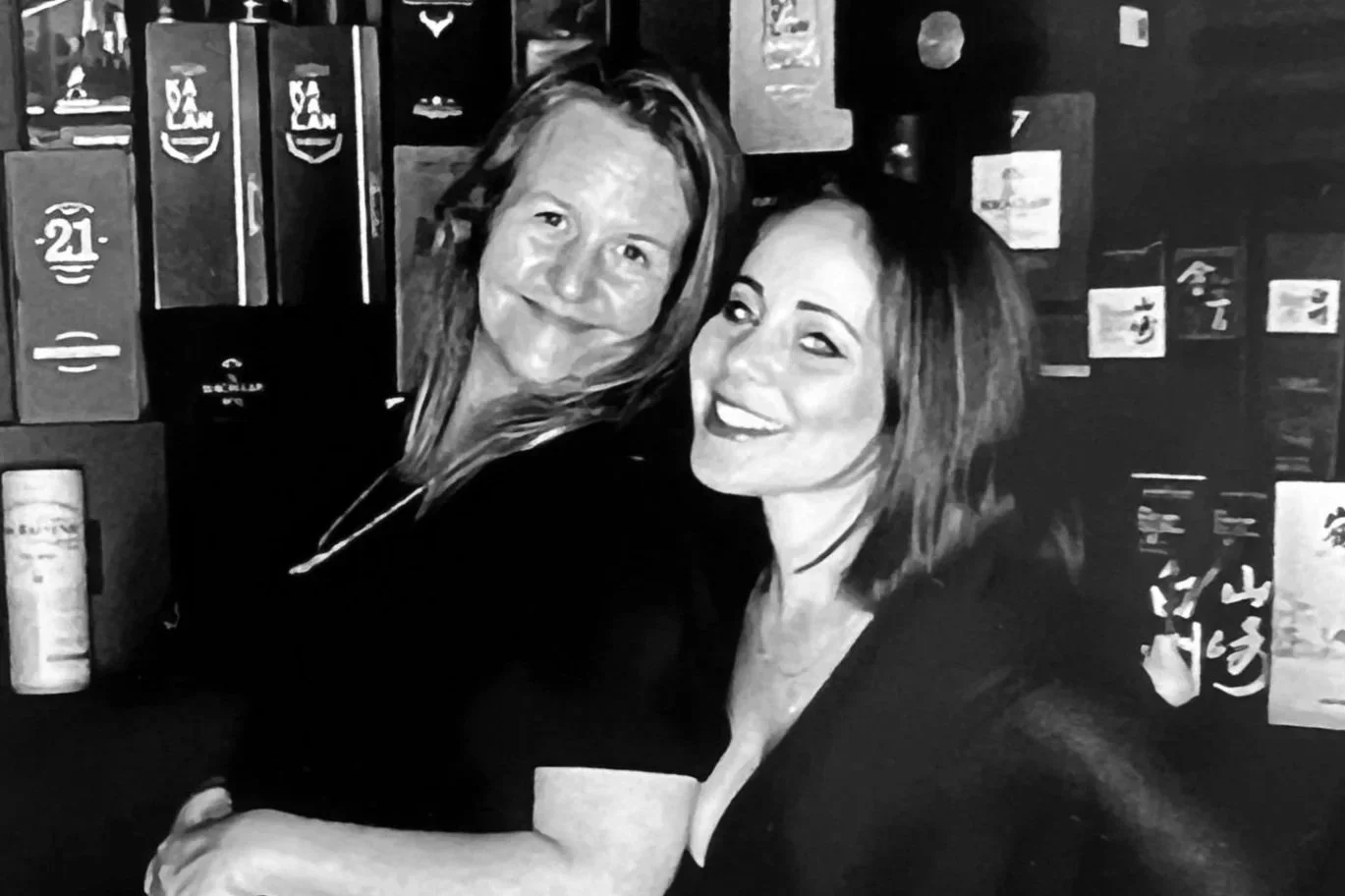 Two women smiling and posing together in front of a wall of liquor bottles.