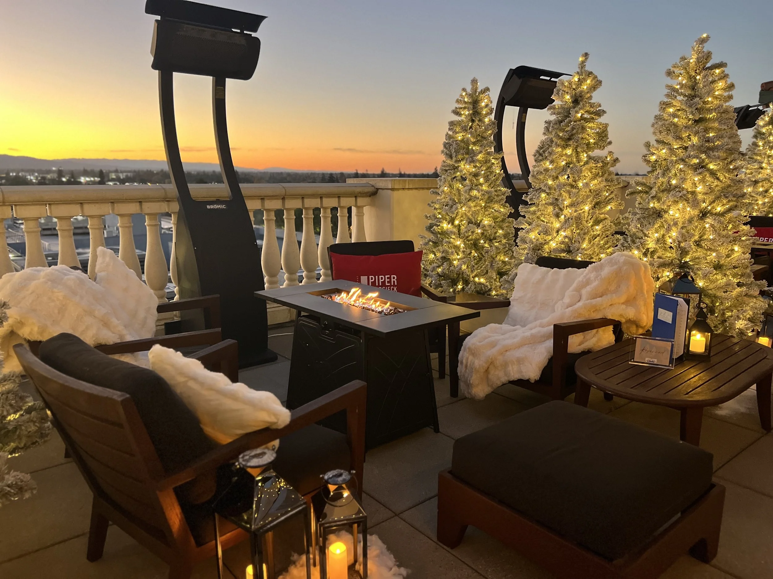 A cozy rooftop terrace, Cielo at Hotel Valencia, decorated for winter with illuminated white Christmas trees, furry throws on chairs, a small fire pit, lanterns, and candles, during sunset. A ReImagined Winter.